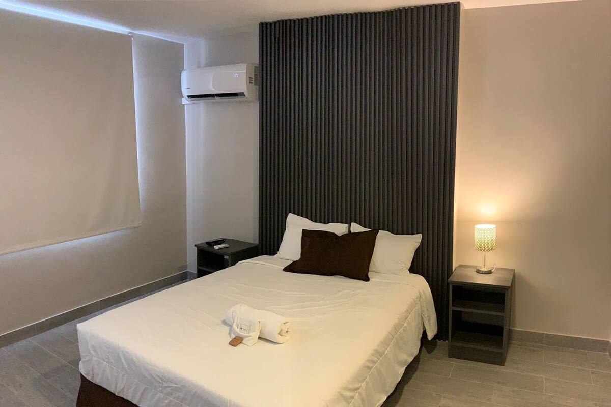 A neatly made bed with white and brown bedding, a bedside lamp, and an air conditioning unit on the wall.