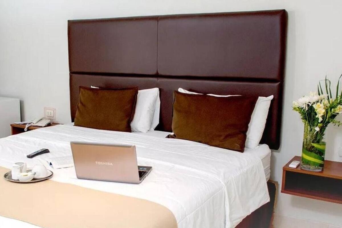 A hotel room with a bed, a laptop, a bedside table with a vase of flowers, and a phone on the nightstand.