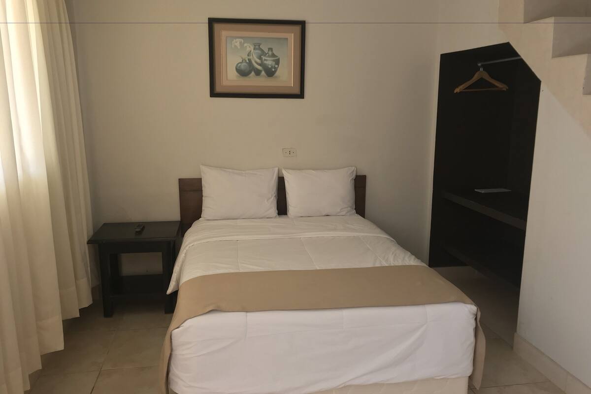 A neatly made bed with white bedding, a small bedside table, a framed picture on the wall, and a hanging coat rack.