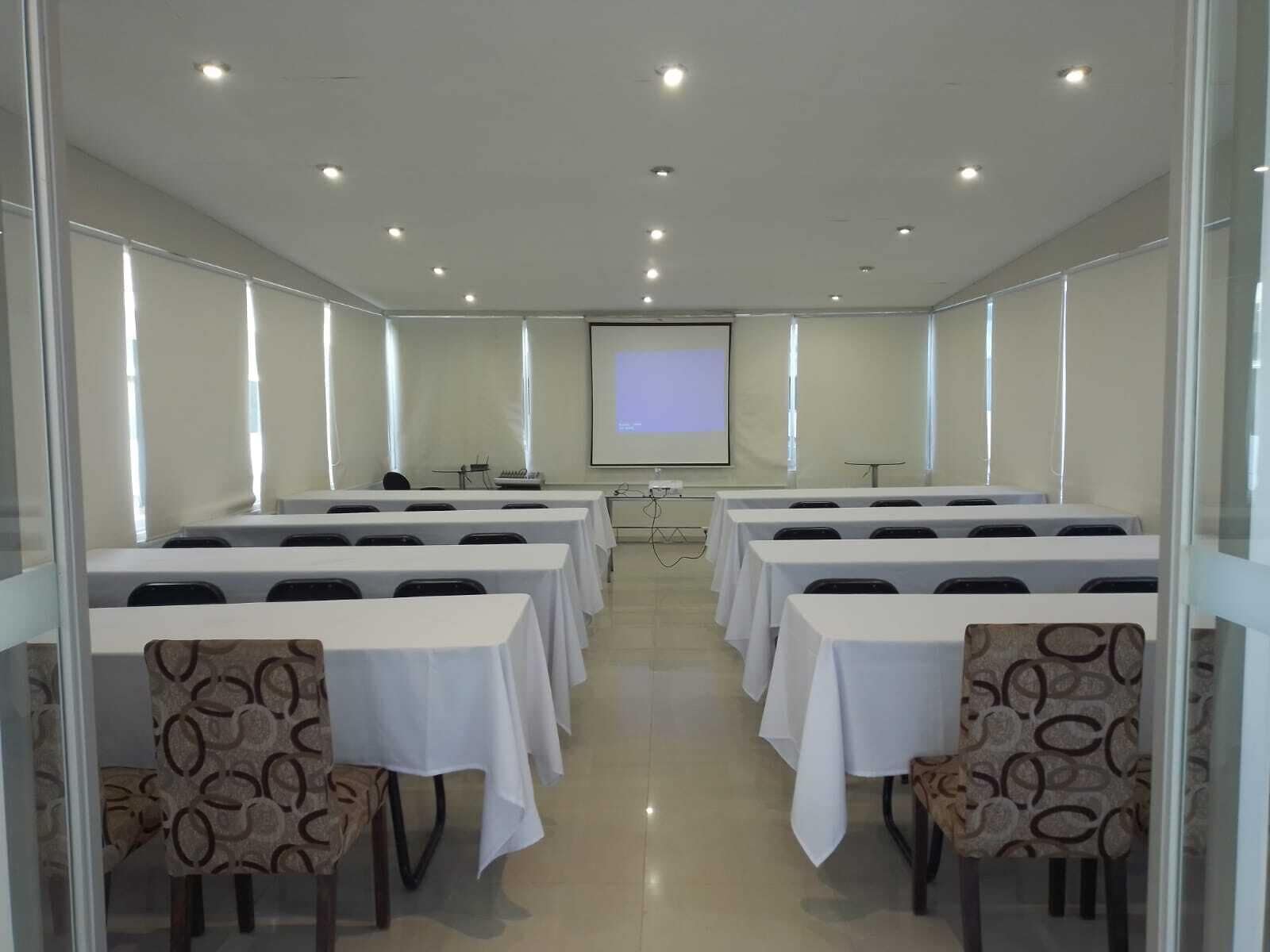 Une salle de conférence équipée de plusieurs tables et chaises, d’un écran de projection et d’un présentoir à l’avant.