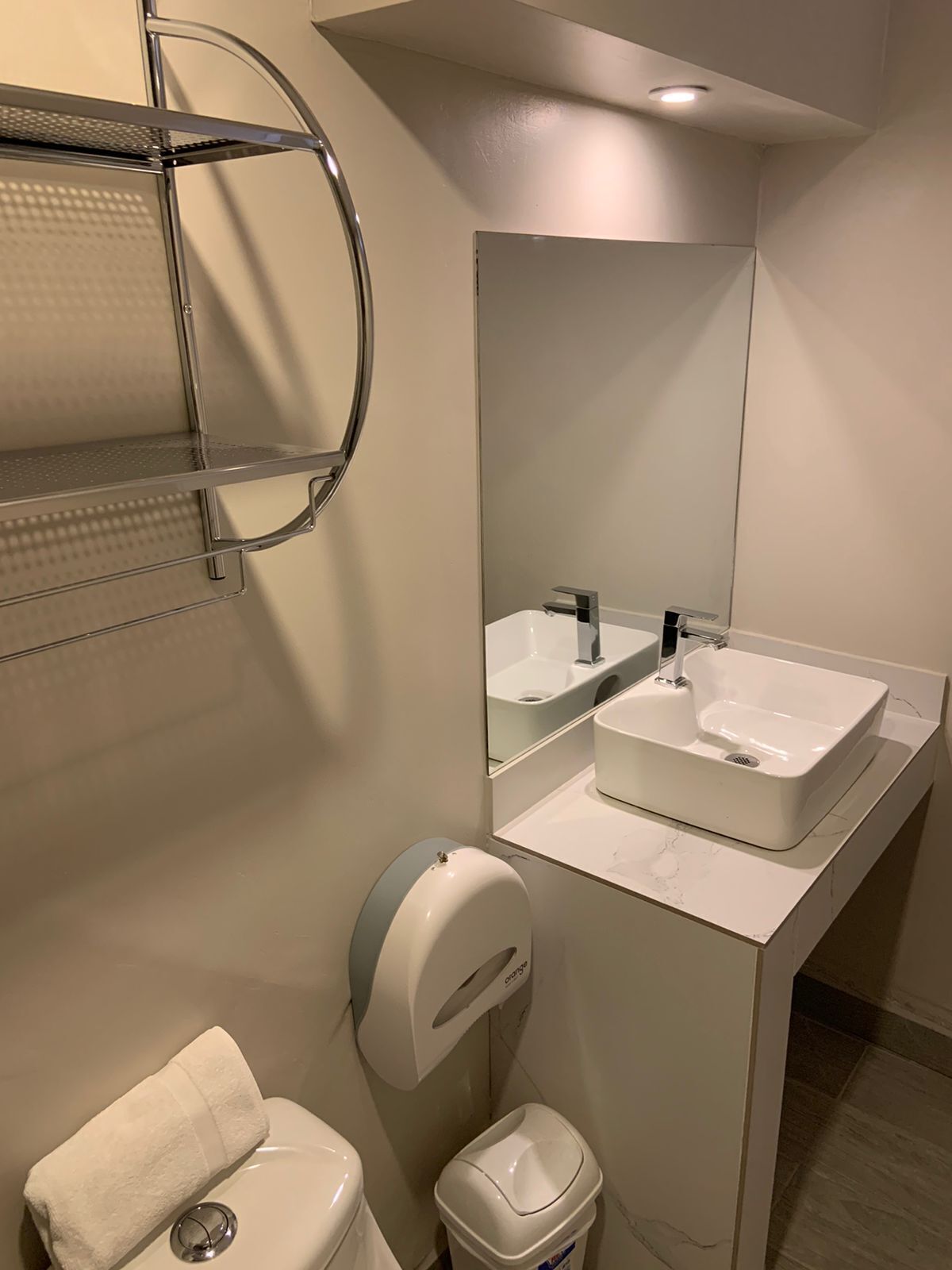 A modern bathroom with a white sink, a mirror, a towel dispenser, and a toilet.