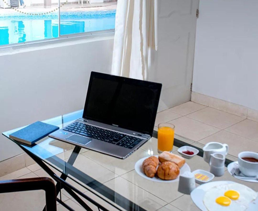 Um laptop sobre uma mesa de vidro com itens de café da manhã, vista para a piscina e o texto 'TRABAJA SEGURO' na parte superior.
