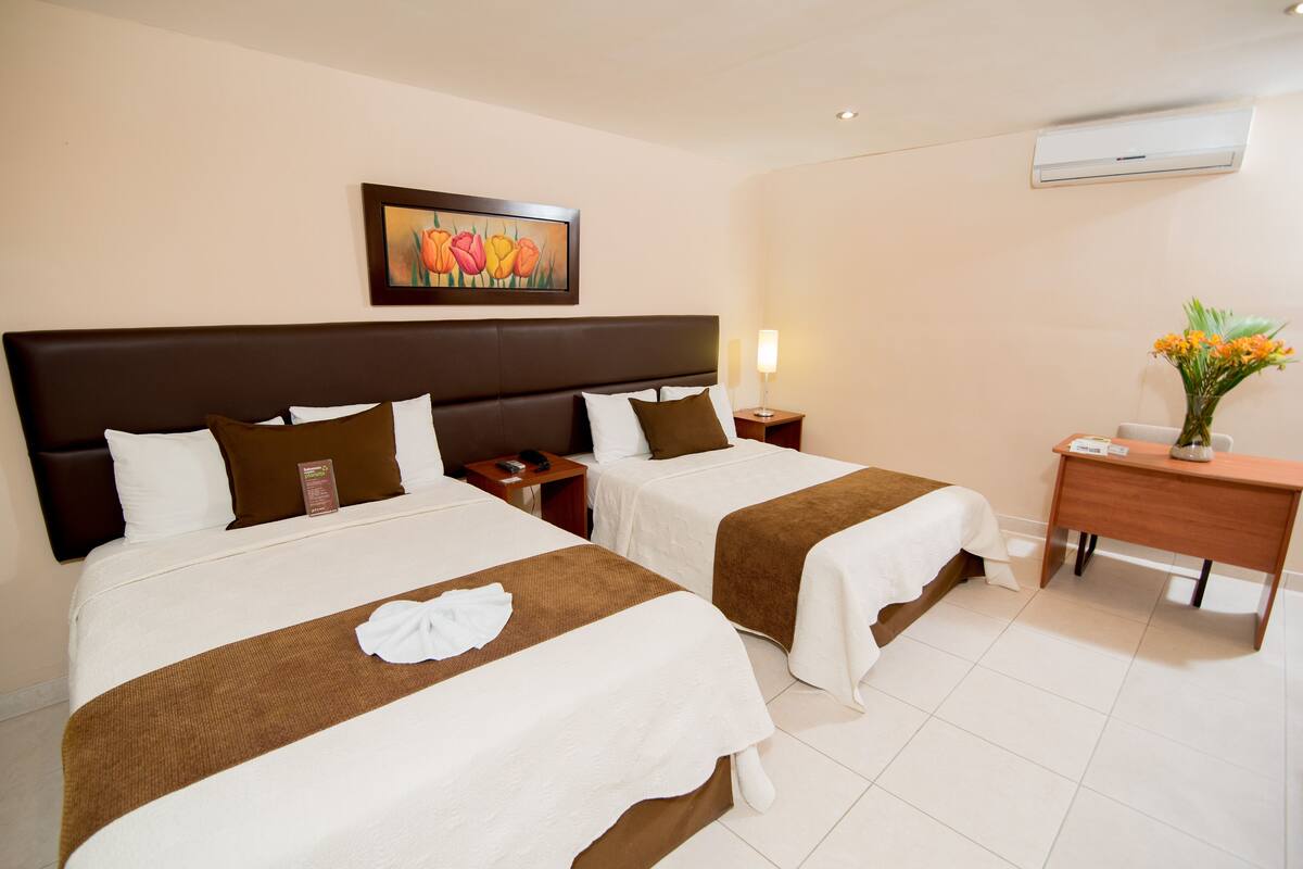 Quarto de hotel com duas camas, uma escrivaninha, uma cadeira, uma mesa de cabeceira, um abajur e um quadro na parede.