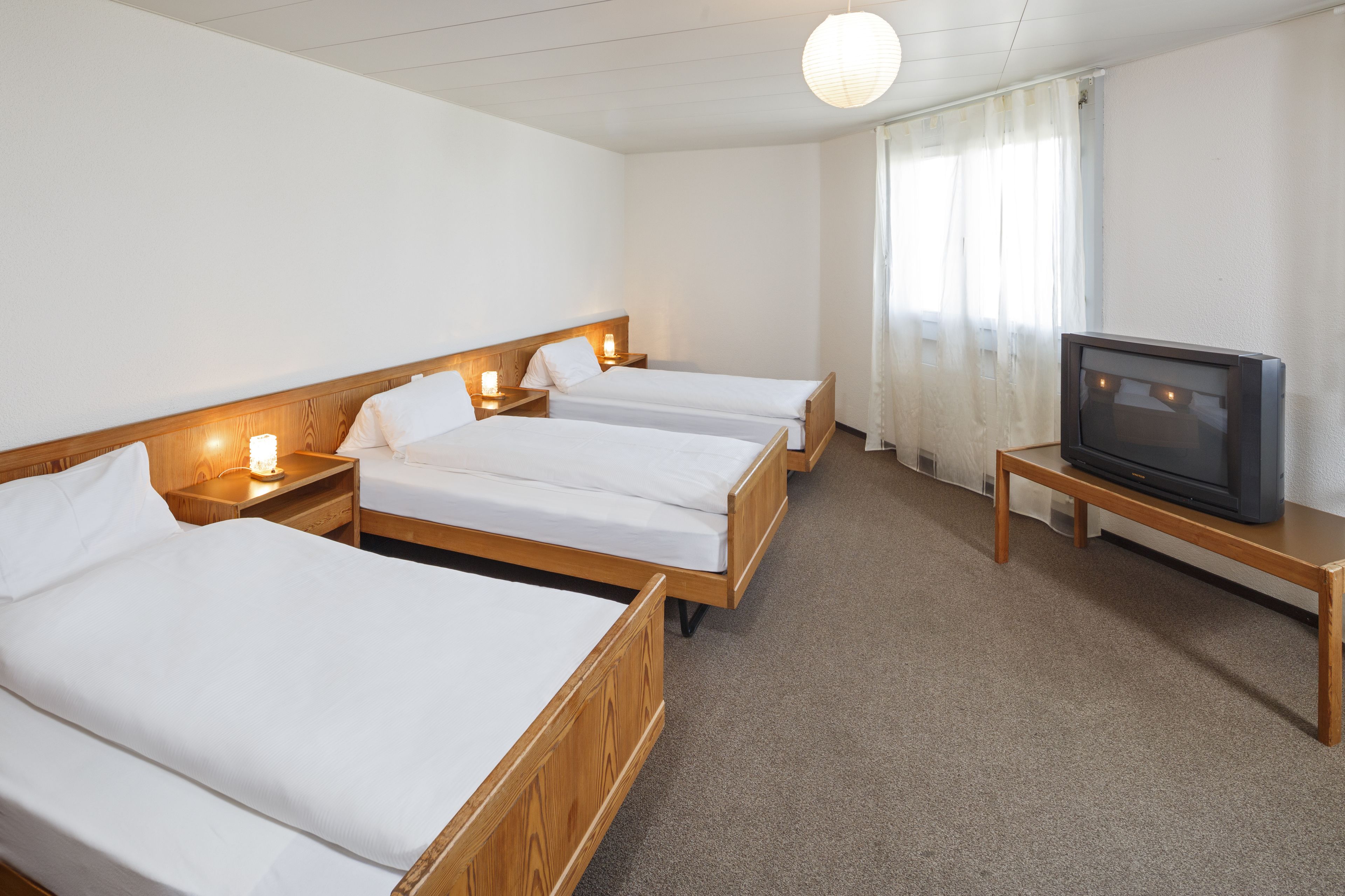 A hotel room with three wooden beds, a television on a wooden stand, and a window with curtains.