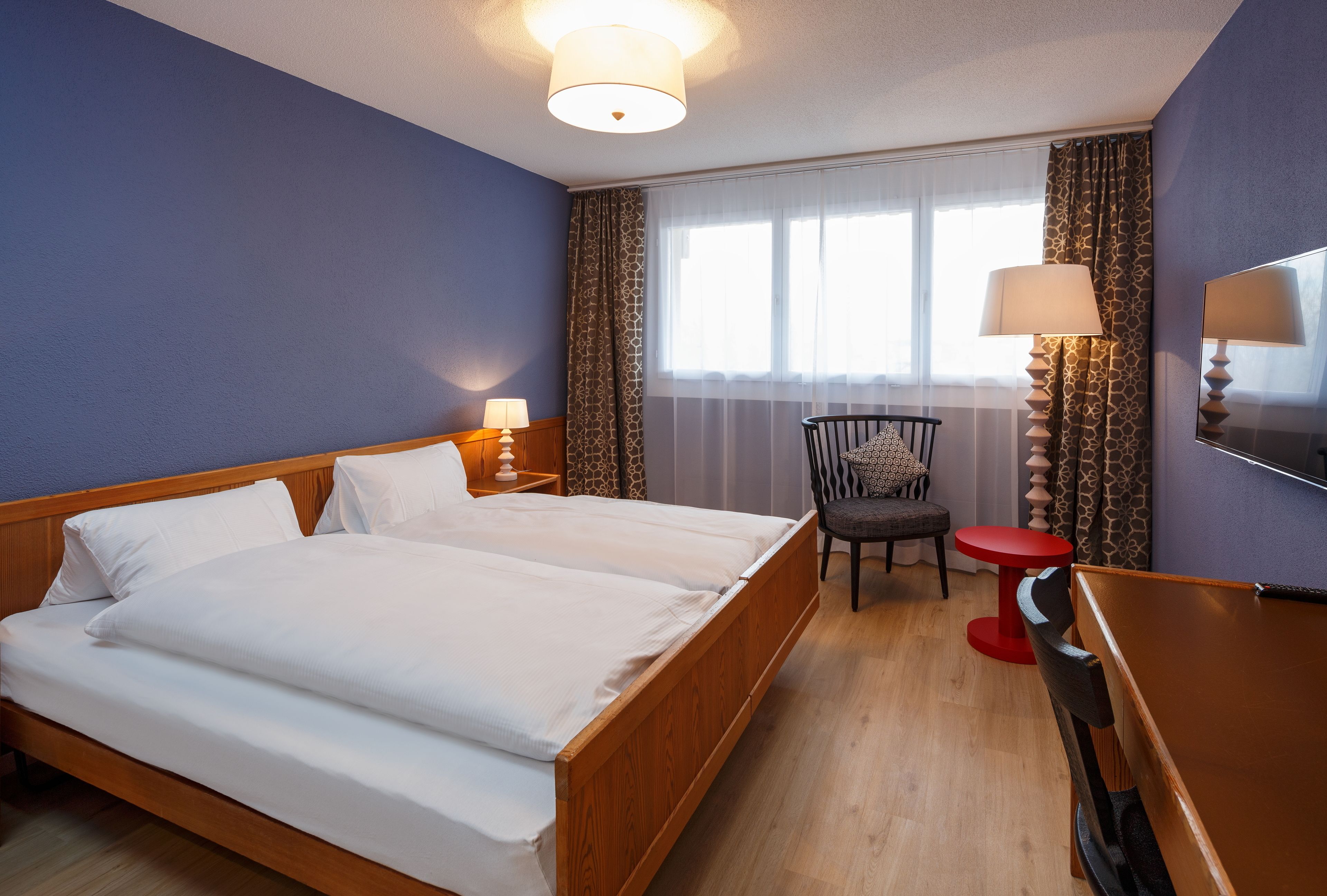 Une chambre d’hôtel avec un lit double, un bureau, une chaise, une télévision et une fenêtre avec des rideaux.