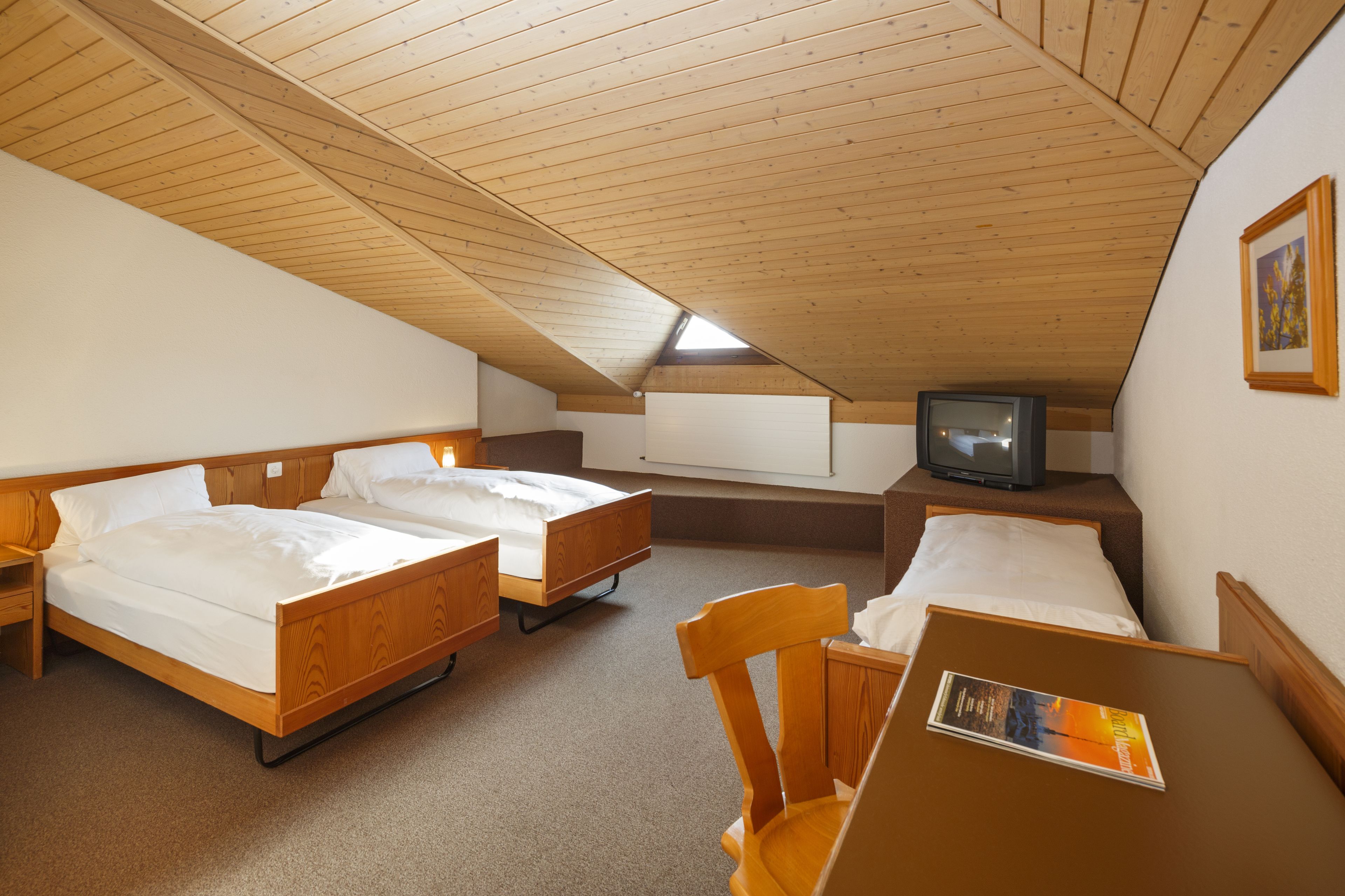 Quarto com duas camas de madeira, uma televisão sobre um suporte, uma cadeira de madeira e uma pequena mesa com uma revista.