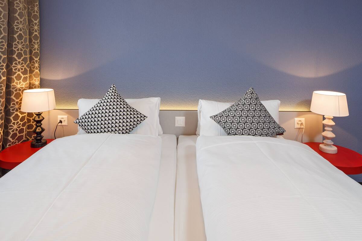 Een hotelkamer met twee bedden, gekleurde kussens, rode nachtkastjes en lampen.