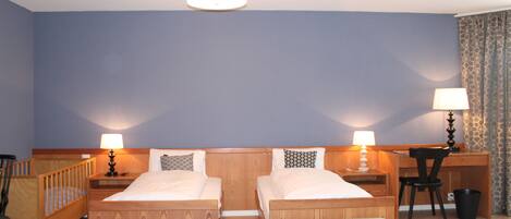A room with two wooden beds, a desk, a chair, a small table, a lamp, and a trash bin.
