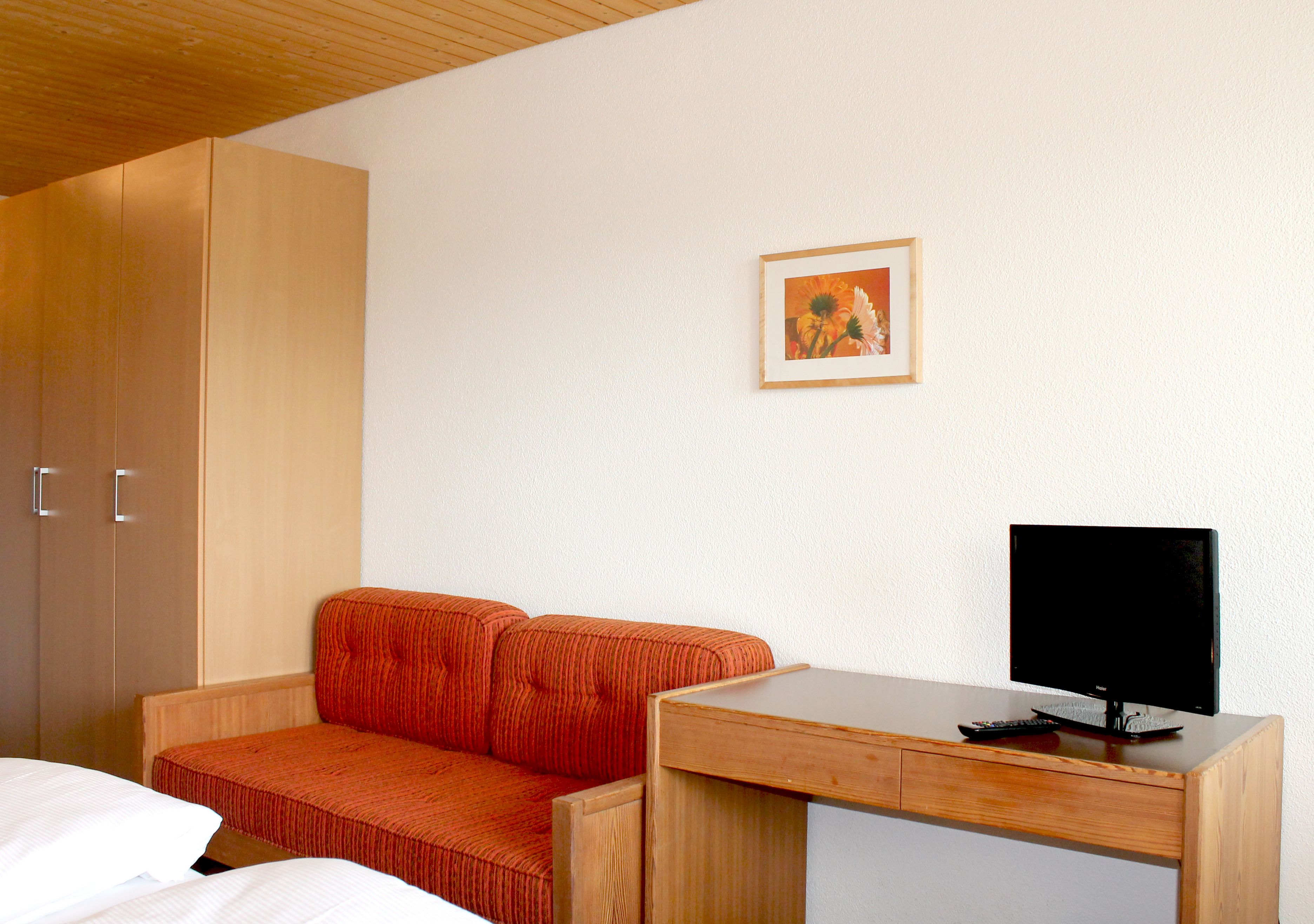 A hotel room with a bed, a wooden desk with a TV, a framed picture on the wall, and a wardrobe.