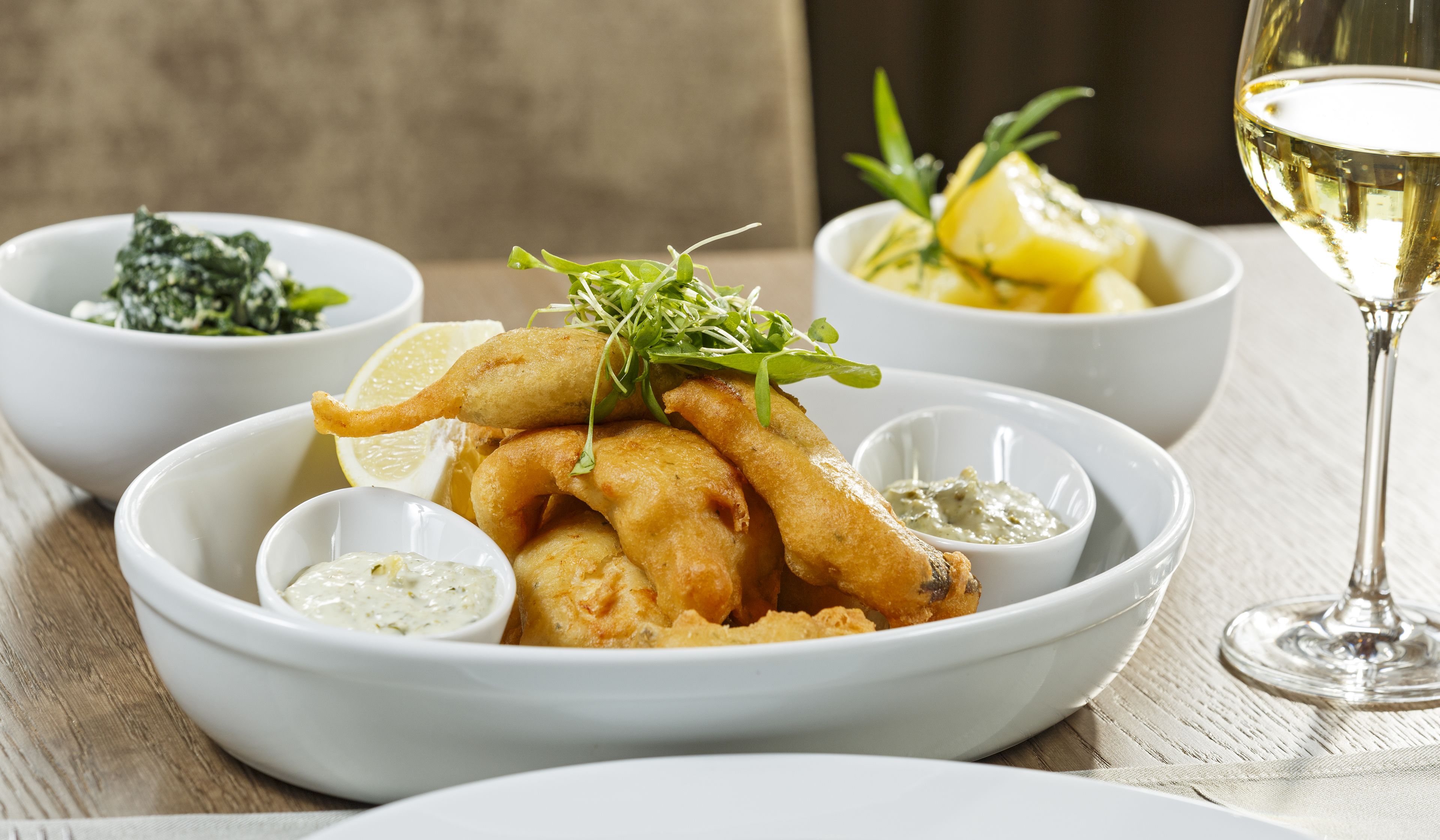 Un plato de pescado frito con cuartos de limón y verduras, acompañado de recipientes pequeños con salsa tártara y una copa de vino blanco.