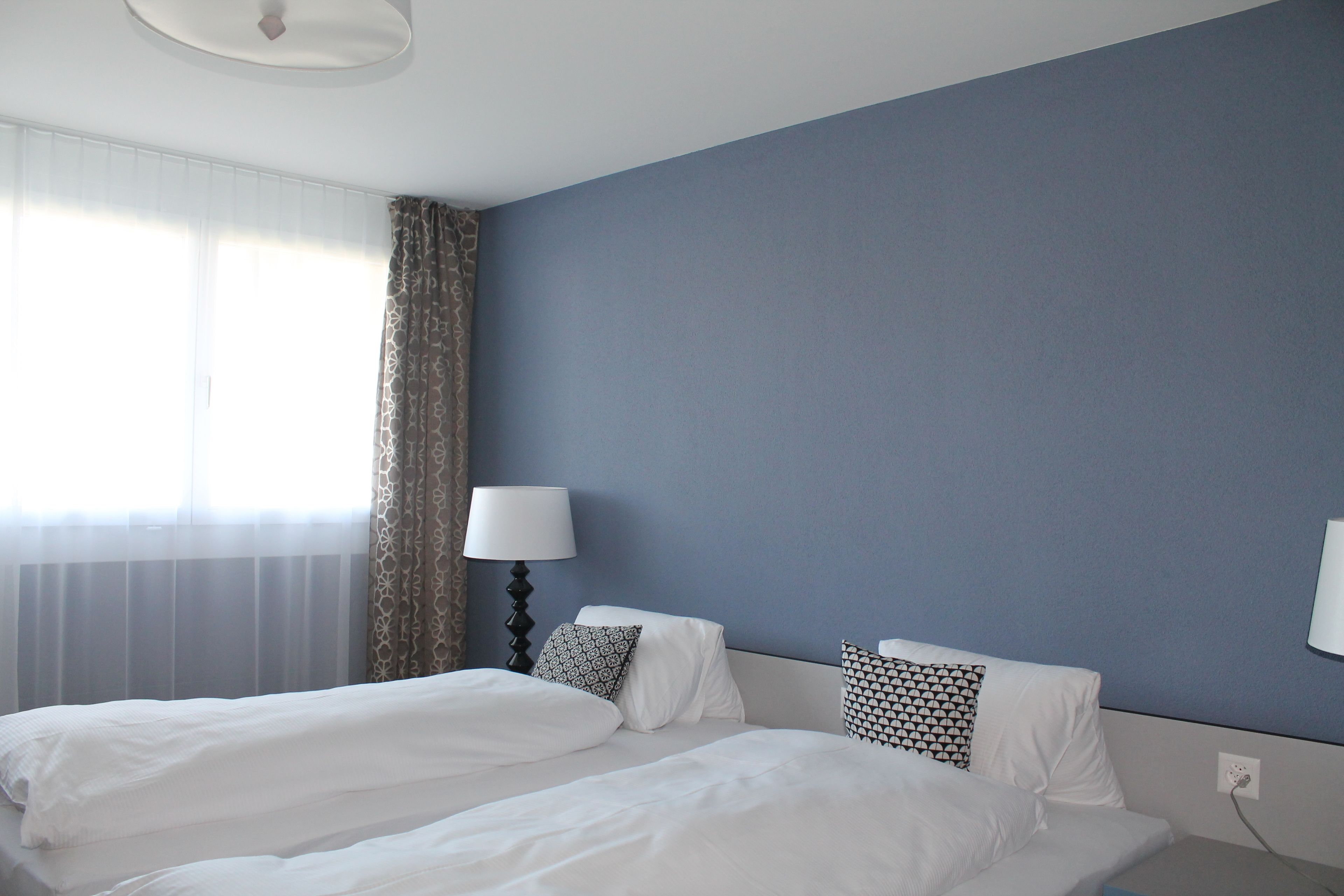 A hotel room with a large bed, two pillows, a bedside lamp, and a window with sheer curtains.