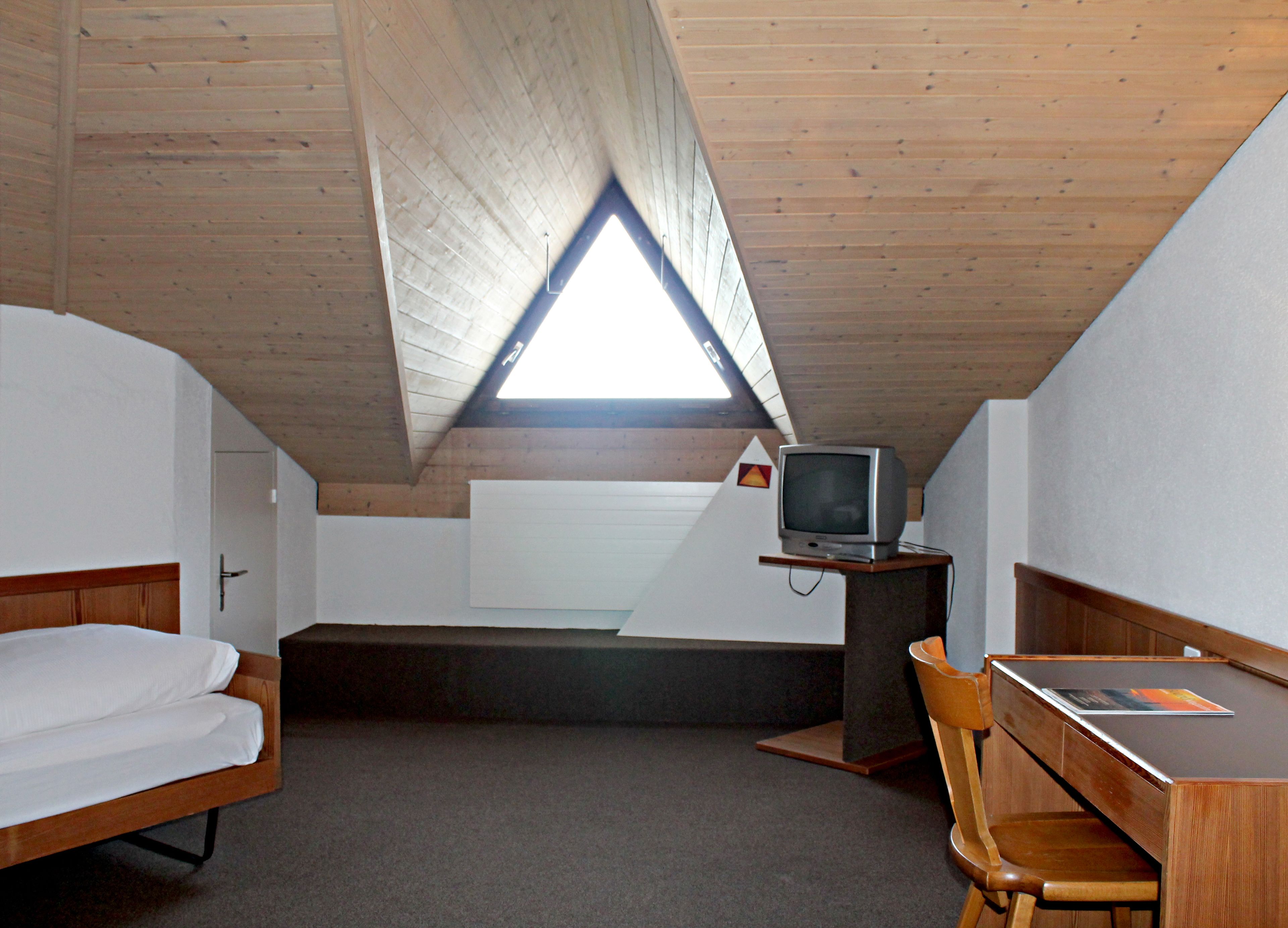 Een kleine, driehoekige zolderkamer met houten plafond, een bed, een bureau en een televisie.