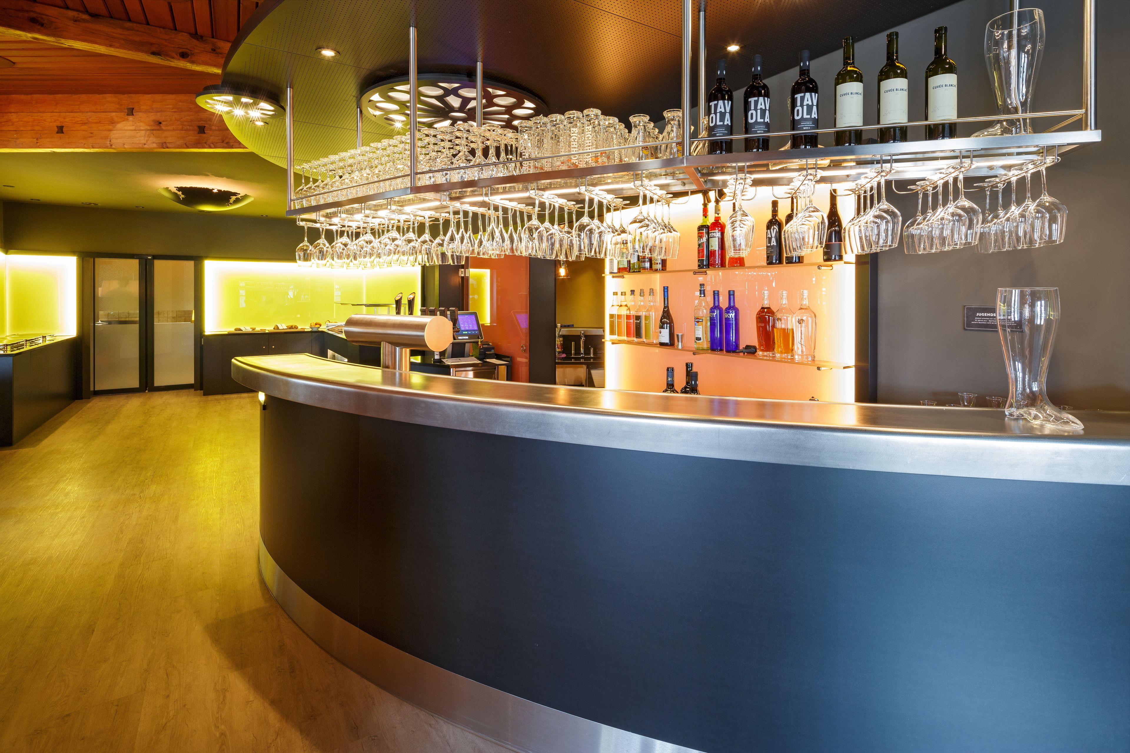 Un espace bar doté d’un comptoir incurvé, d’étagères remplies de bouteilles et de verres, et d’un plafond en bois.