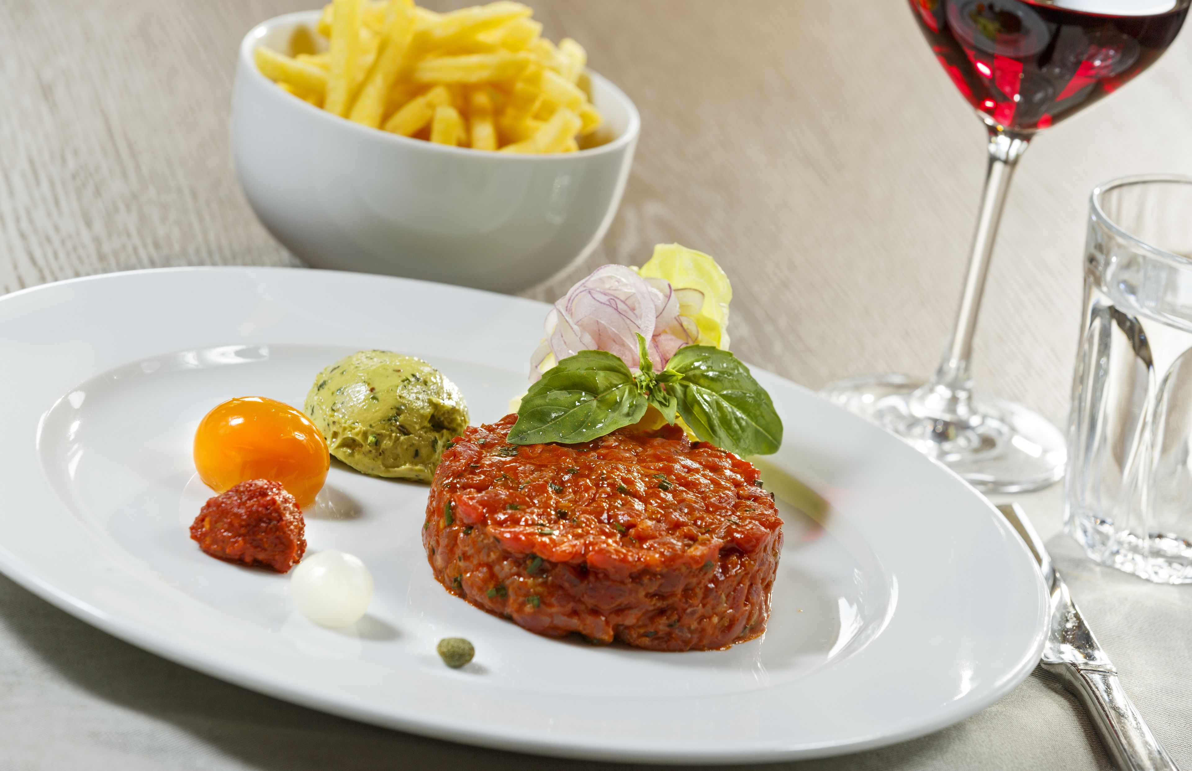 Un plat amb tartare de vedella, un bol de patates fregides i un got de vi vermell.