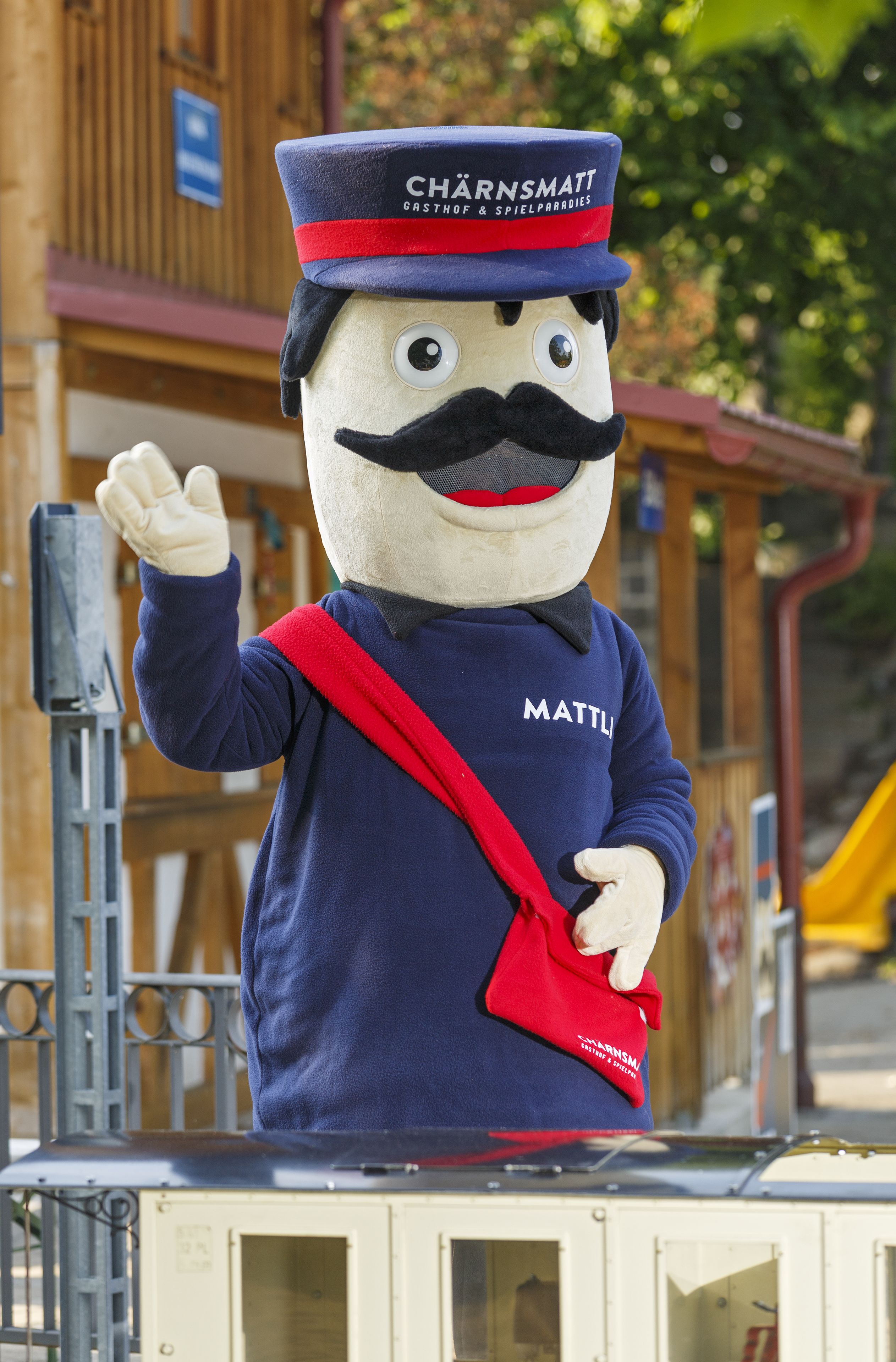 A mascot with a blue uniform and red sash, standing on a platform with a wooden structure in the background.