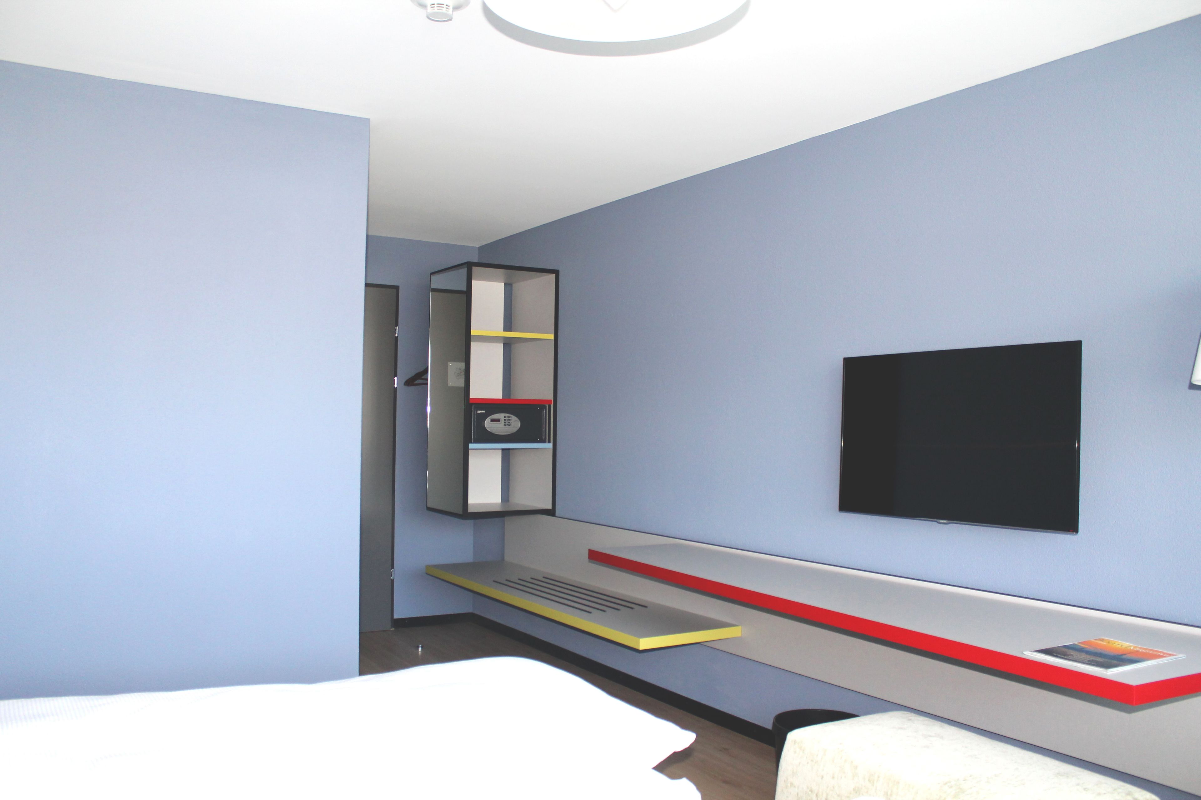 Ein modernes Hotelzimmer mit einem Bett, einem an der Wand befestigten Fernseher, einer eingebauten Ablagefläche mit rotem Akzent und einer kleinen Schreibtischzeile.
