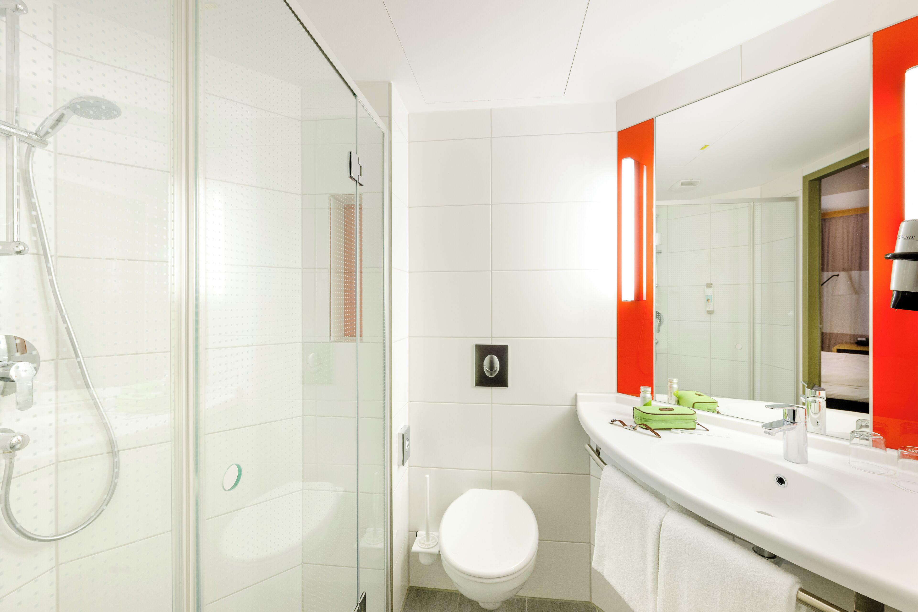 Ein modernes Badezimmer mit Duschkabine aus Glas, einer weißen Toilette und einem Waschtisch mit großem Spiegel und roten Akzenten.