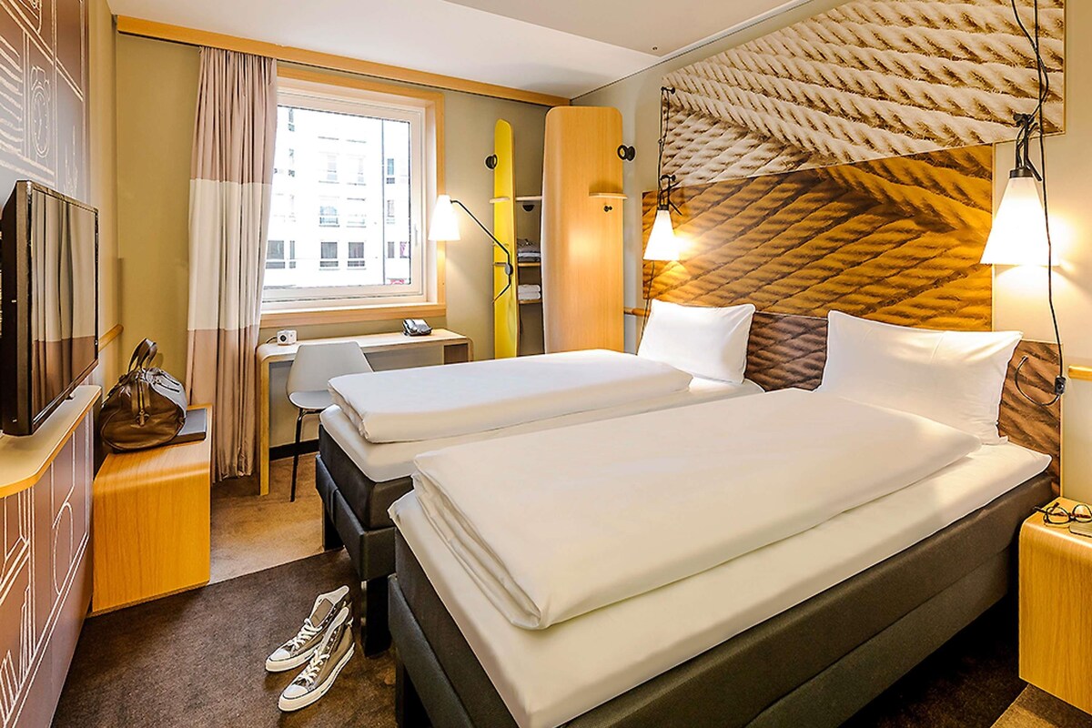 A hotel room with two beds, a desk, a chair, a TV, and a window with curtains.