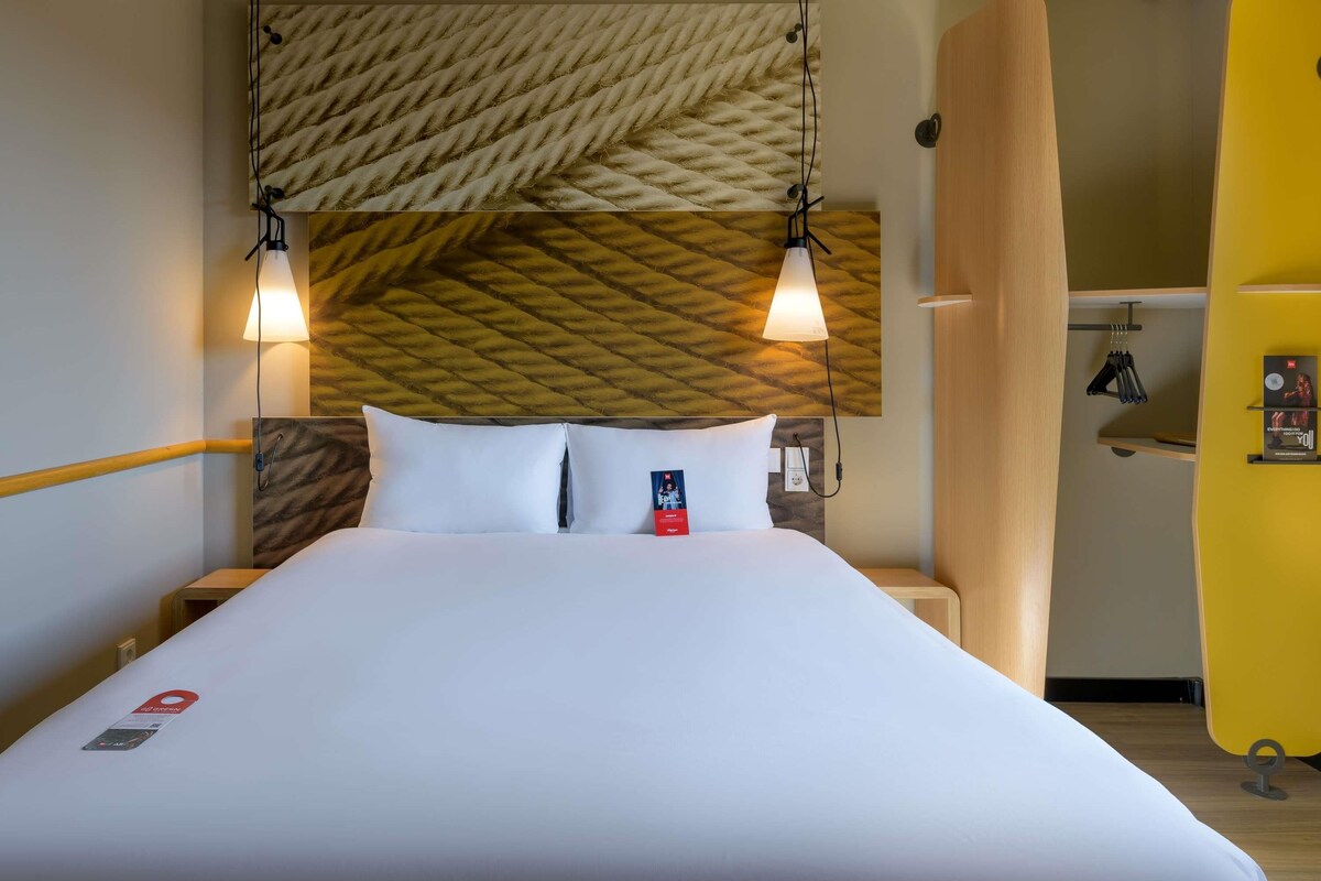 A hotel room with a bed, two bedside lamps, a wooden headboard, a yellow wall, and a small table with items on it.