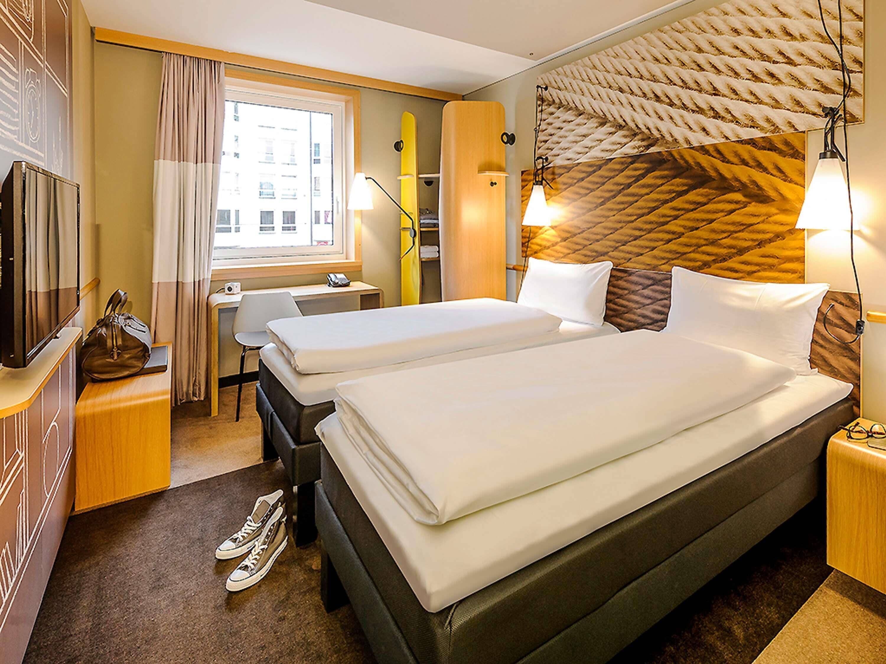 Ein Hotelzimmer mit zwei Betten, einem Schreibtisch, einem Stuhl, einem Fernseher und einem Fenster mit Vorhängen.