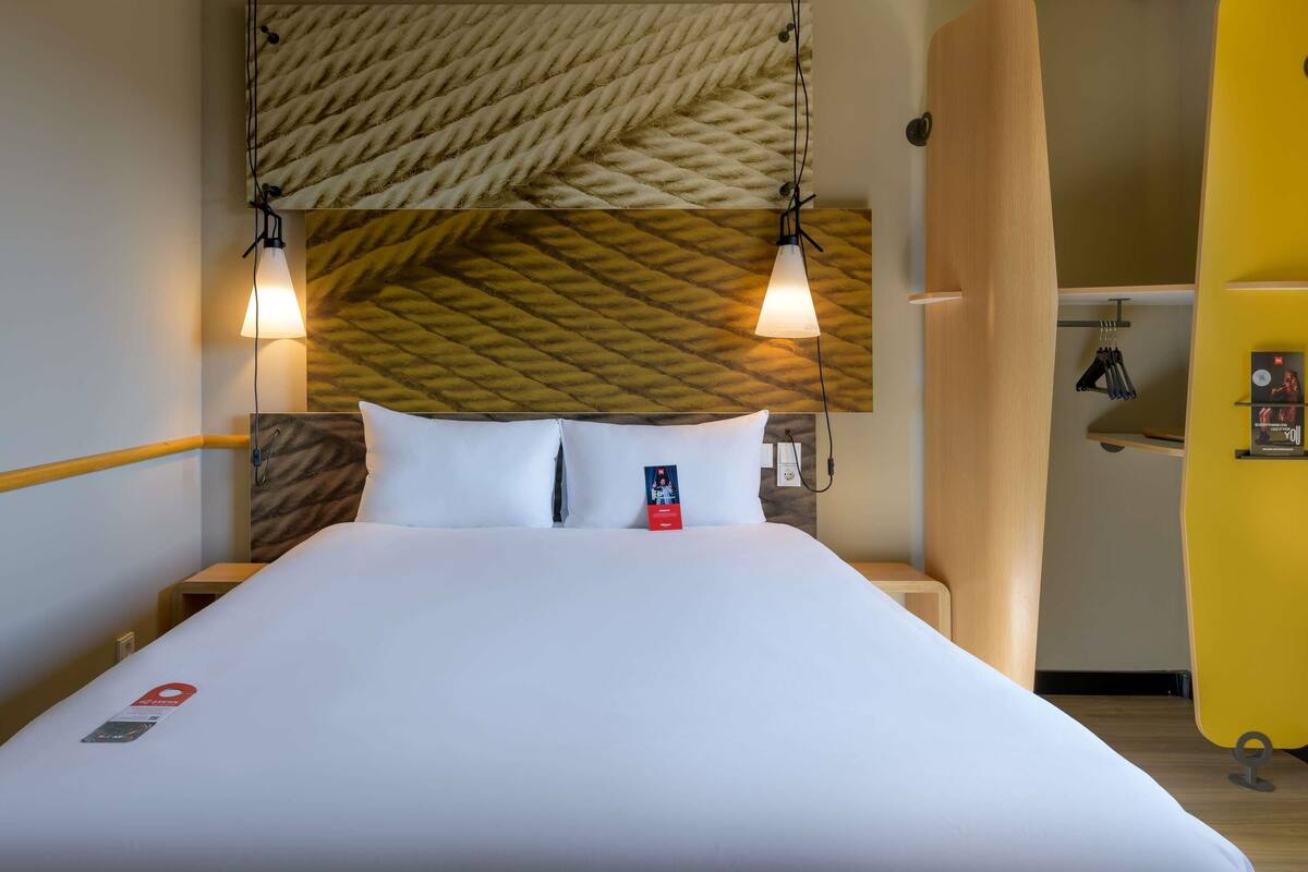 A hotel room with a bed, two bedside lamps, a wooden headboard, a yellow wall, and a small table with items on it.
