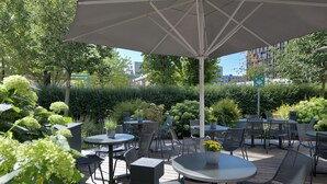 An outdoor patio with round tables and chairs, surrounded by greenery and a large umbrella. - Ibis Muenchen City Arnulfpark (Munich)