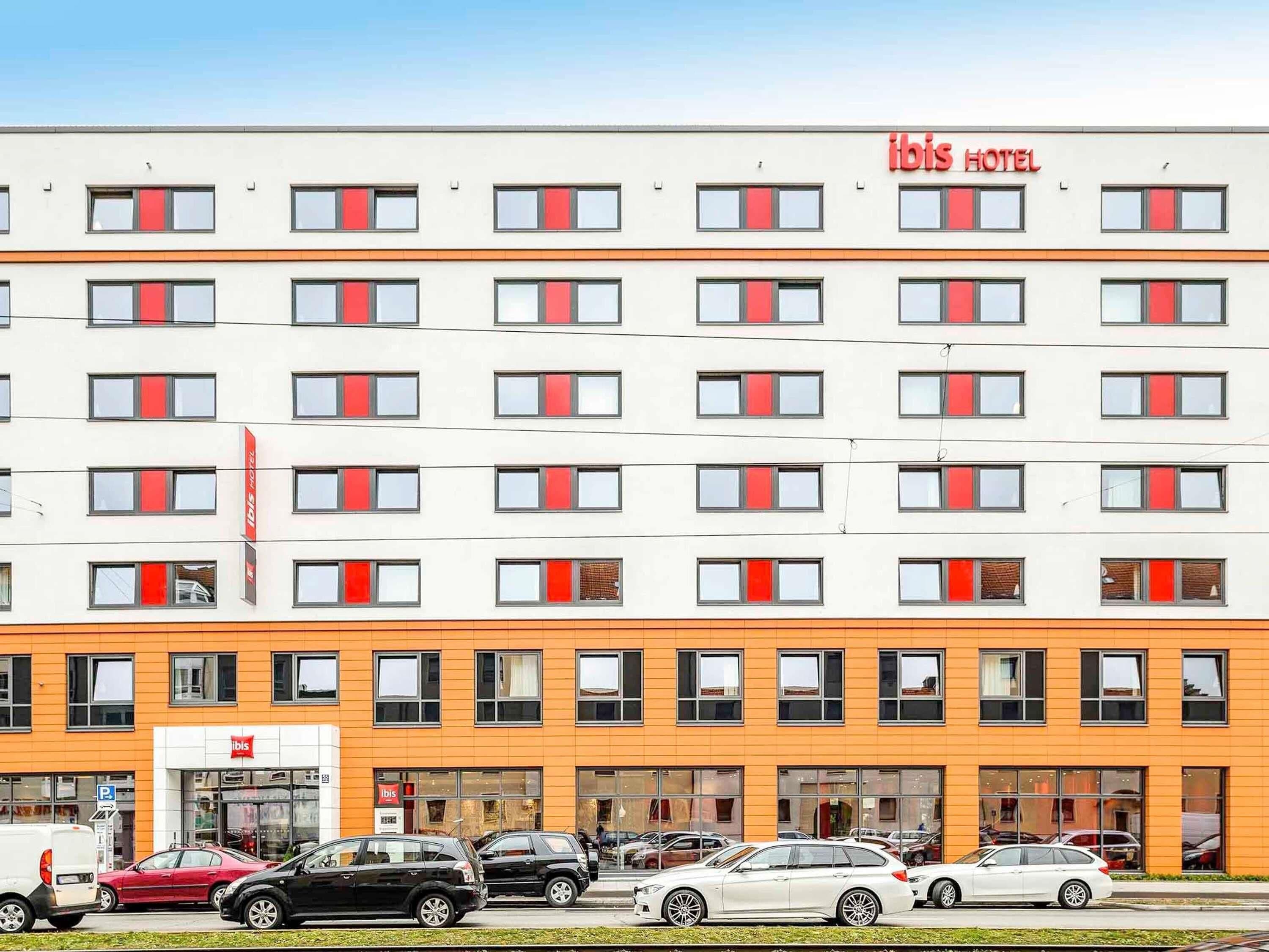 Un hotel de varios pisos con fachada moderna, que cuenta con una serie de ventanas con cortinas rojas y un llamativo letrero 'ibis HOTEL'.