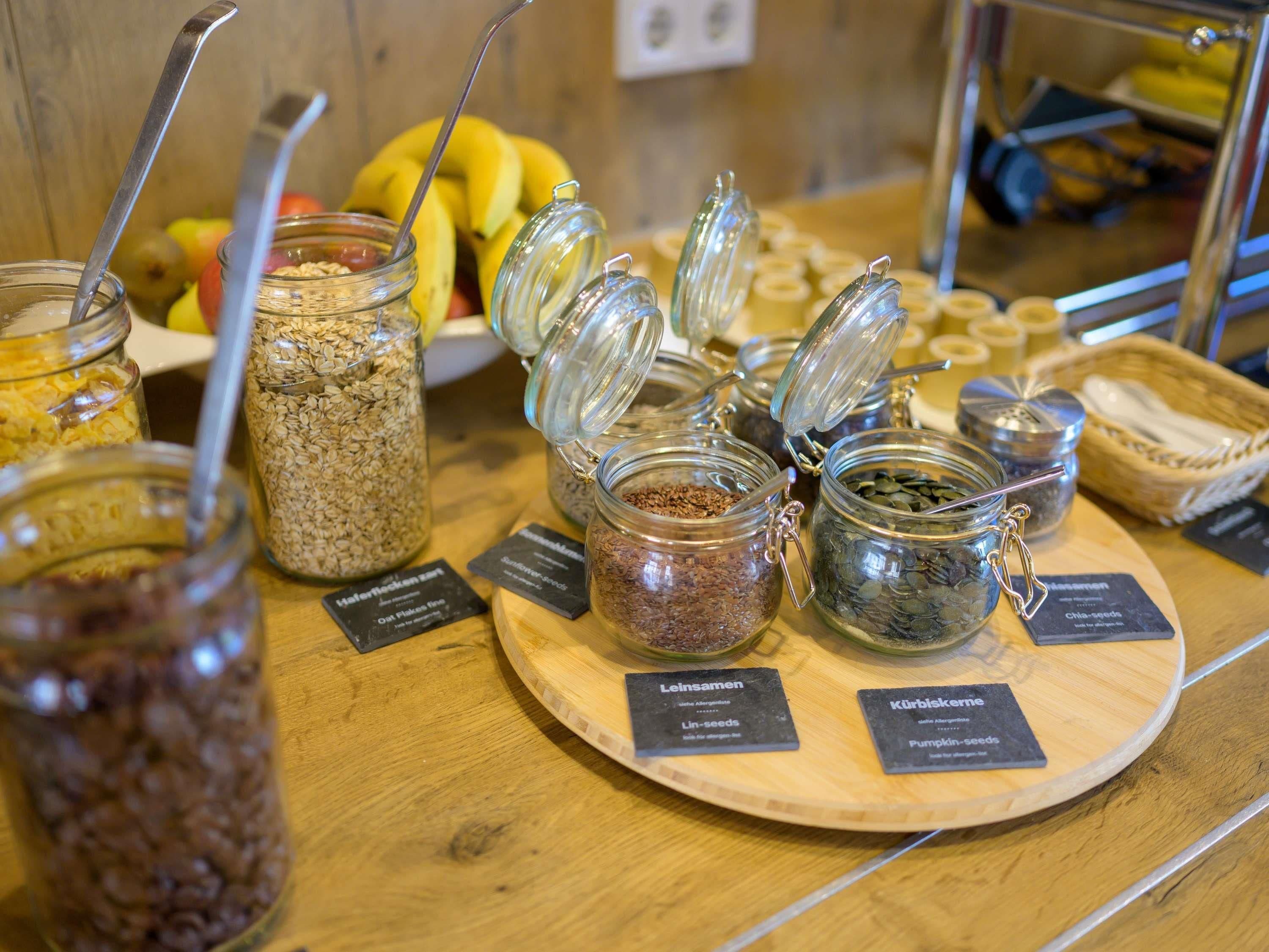 A variety of food items in jars with labels, a wooden table, and a fruit bowl in the background.