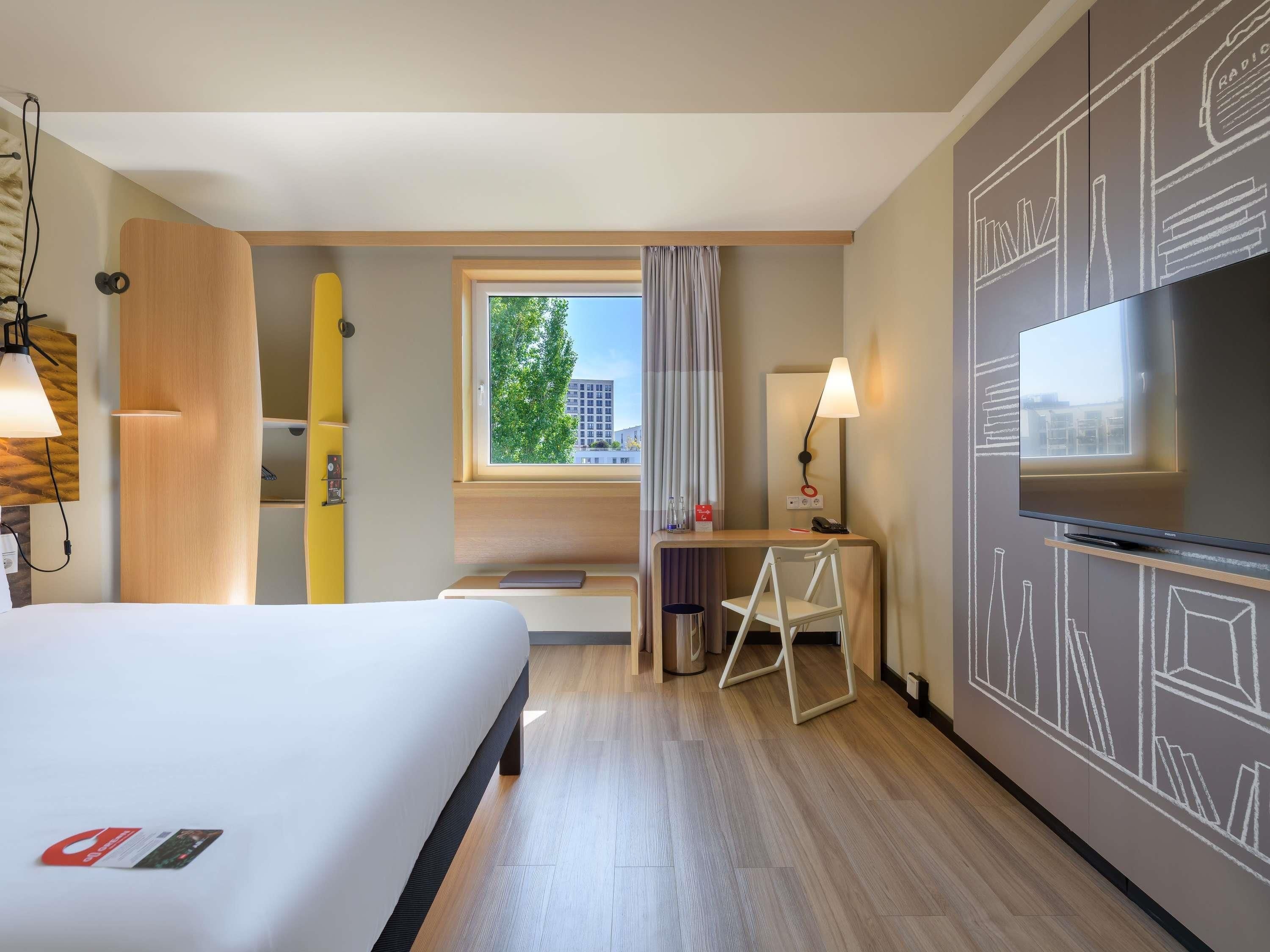 A hotel room with a bed, desk, chair, TV, and a window with a view of buildings.