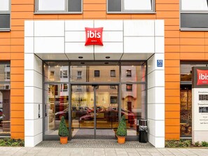 A hotel entrance with a glass facade, two potted plants, and a red sign above the doors. - Ibis Muenchen City Arnulfpark (Munich)