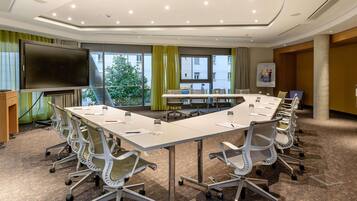 Un salón de conferencias con una larga mesa, sillas, una pantalla de proyector y ventanas amplias con cortinas.