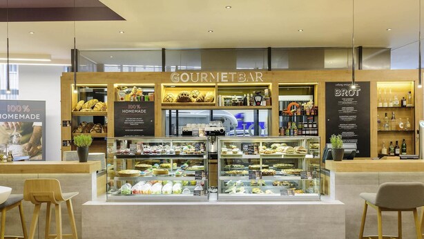 A gourmet bar with a variety of baked goods and sandwiches displayed in a glass case, wooden counters, and a sign indicating 100% homemade products. - Ibis Muenchen City Arnulfpark (Munich)