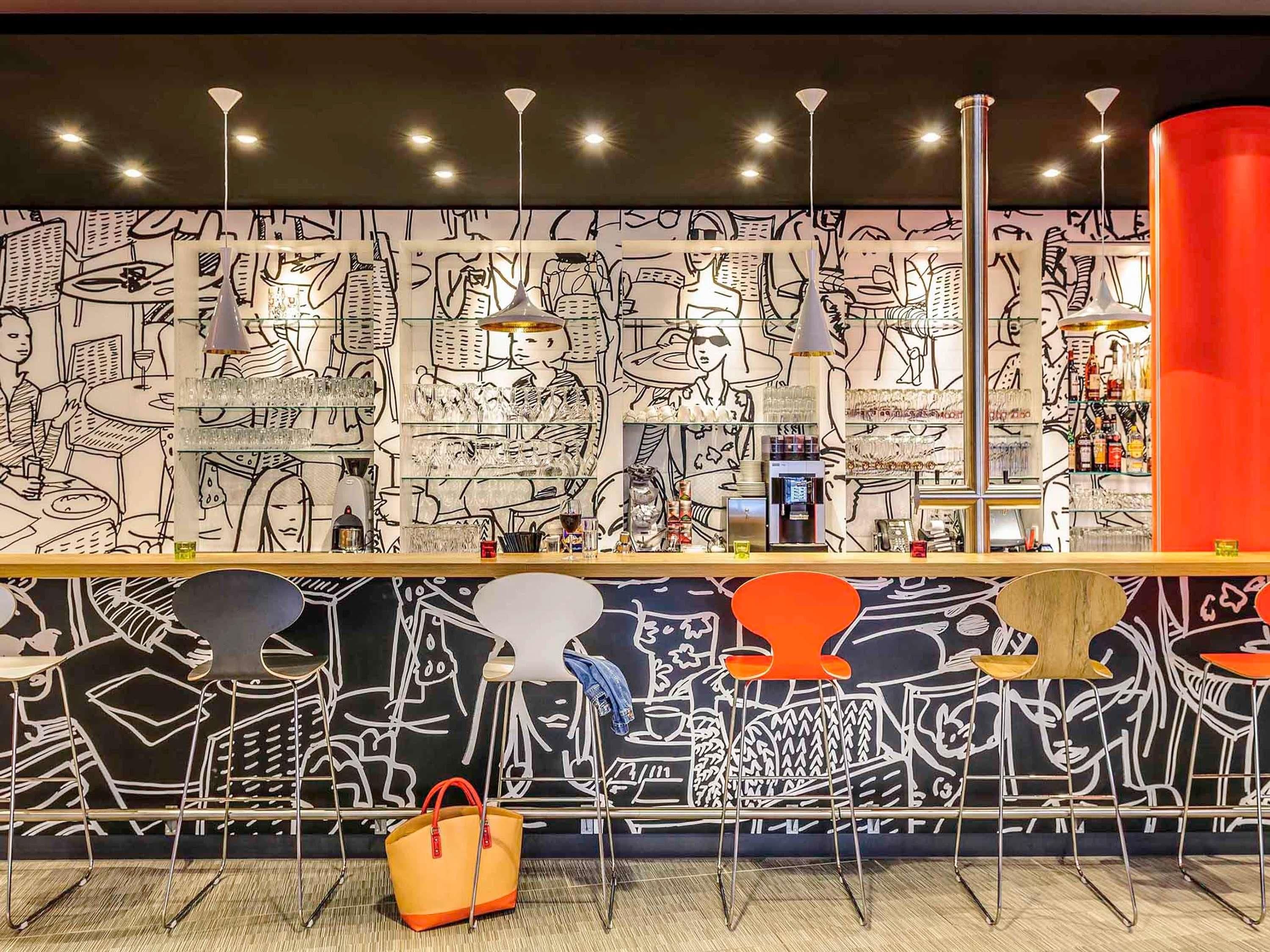 Zona de bar con un mural que muestra a personas comiendo y bebiendo, taburetes y una barra con caja registradora.