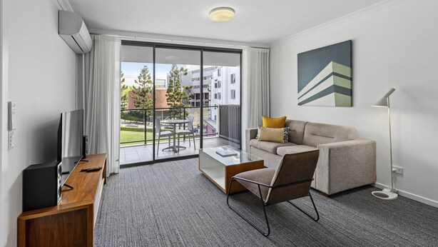 Una habitación de hotel moderna con sofá, una mesa de centro, televisor y balcón con vistas a edificios y árboles.