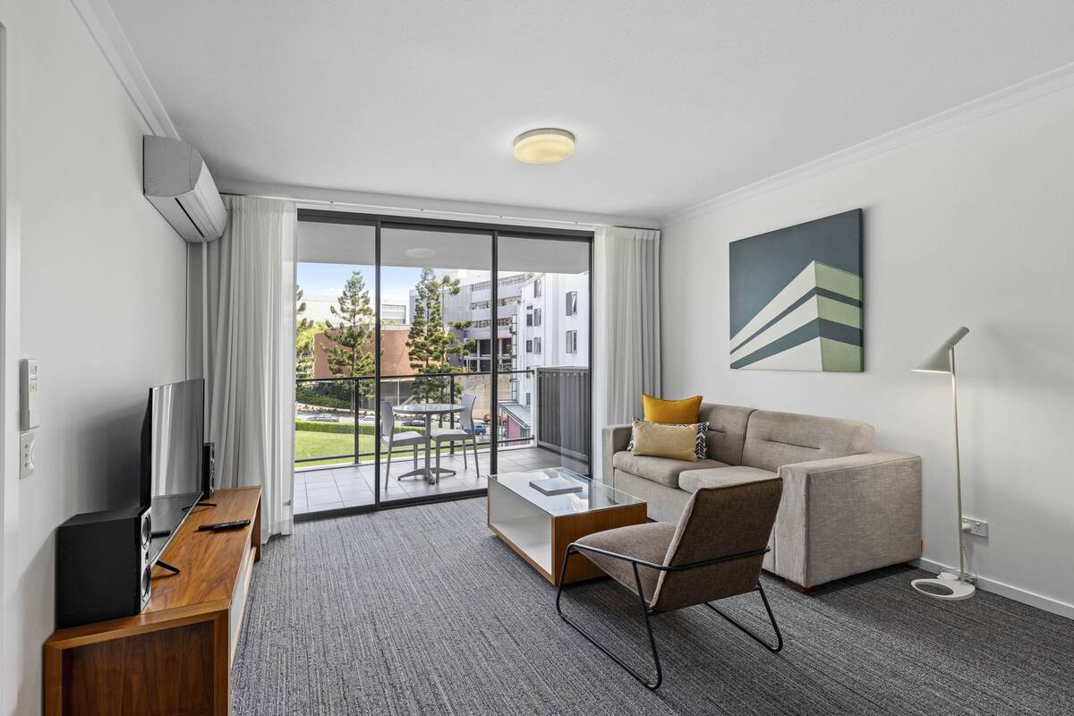 Una habitación de hotel moderna con sofá, una mesa de centro, televisor y balcón con vistas a edificios y árboles.