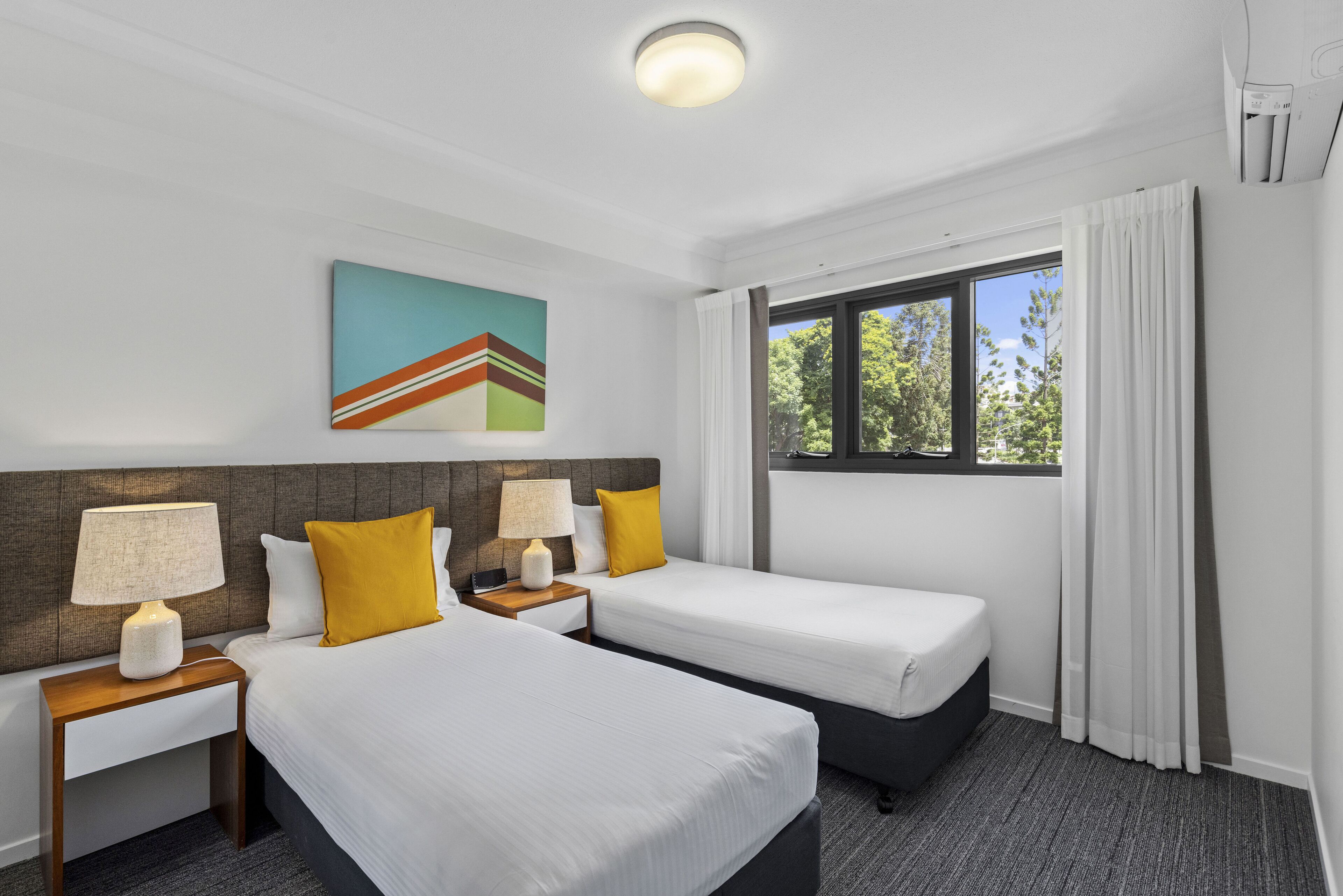 Une chambre d’hôtel avec deux lits, une table de chevet avec une lampe, un tableau au mur et une fenêtre avec des rideaux.
