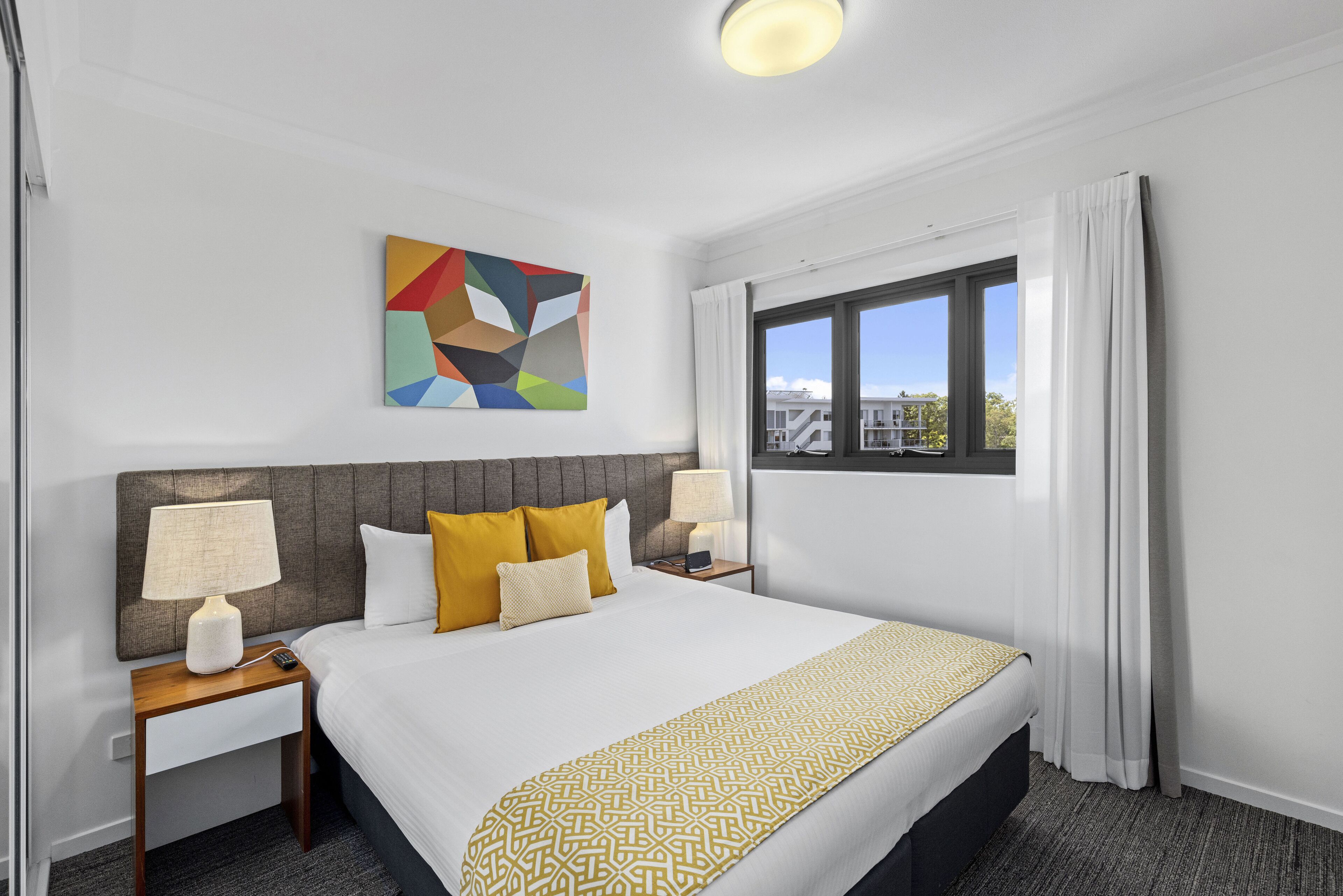 A hotel room with a large bed, two bedside tables with lamps, a colorful abstract painting, and a window with curtains.