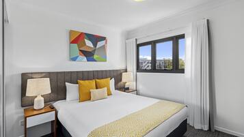 A hotel room with a large bed, two bedside tables with lamps, a colorful abstract painting, and a window with curtains.
