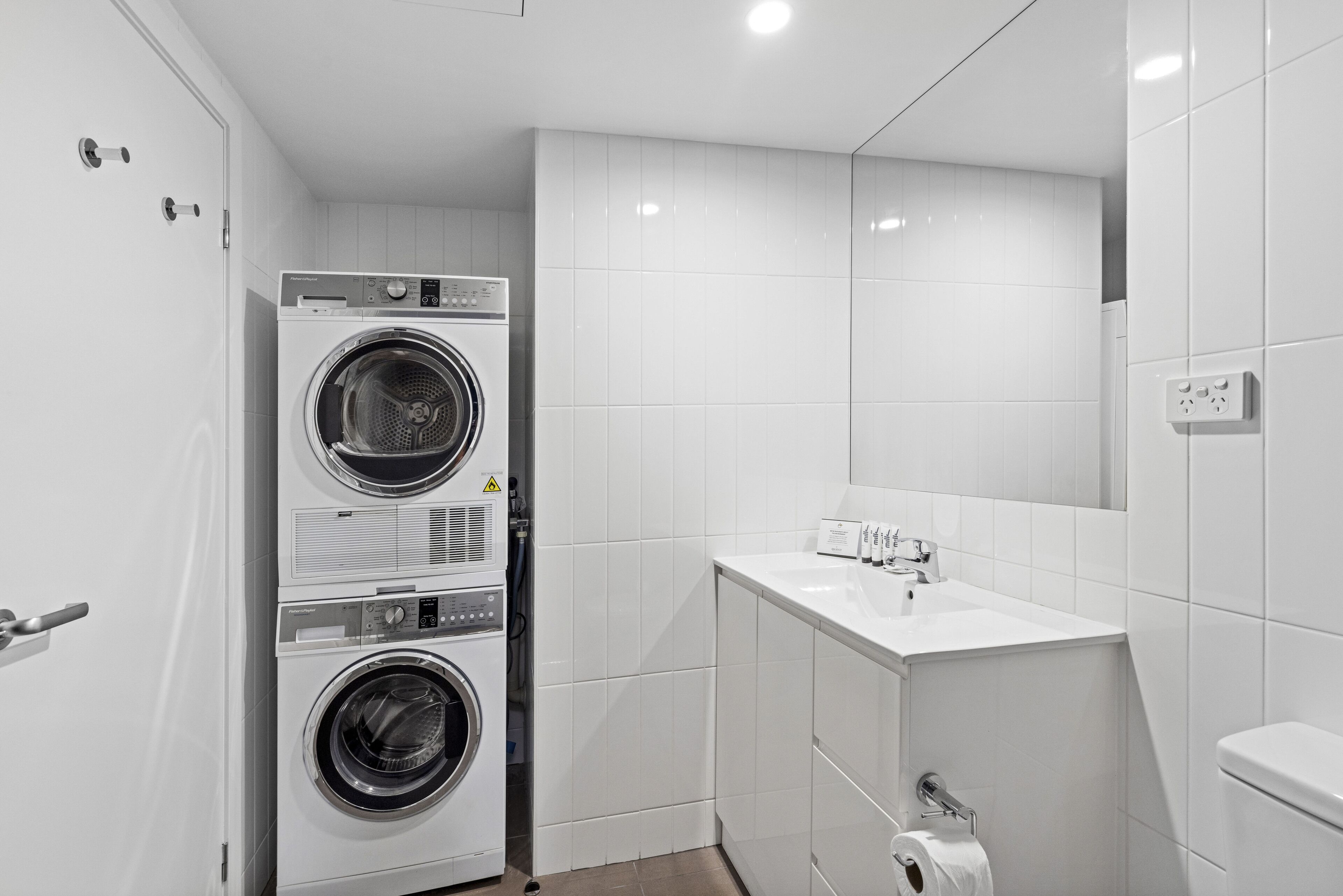 Un bagno compatto e moderno con lavatrice e asciugatrice affiancate, un lavandino bianco e un water.