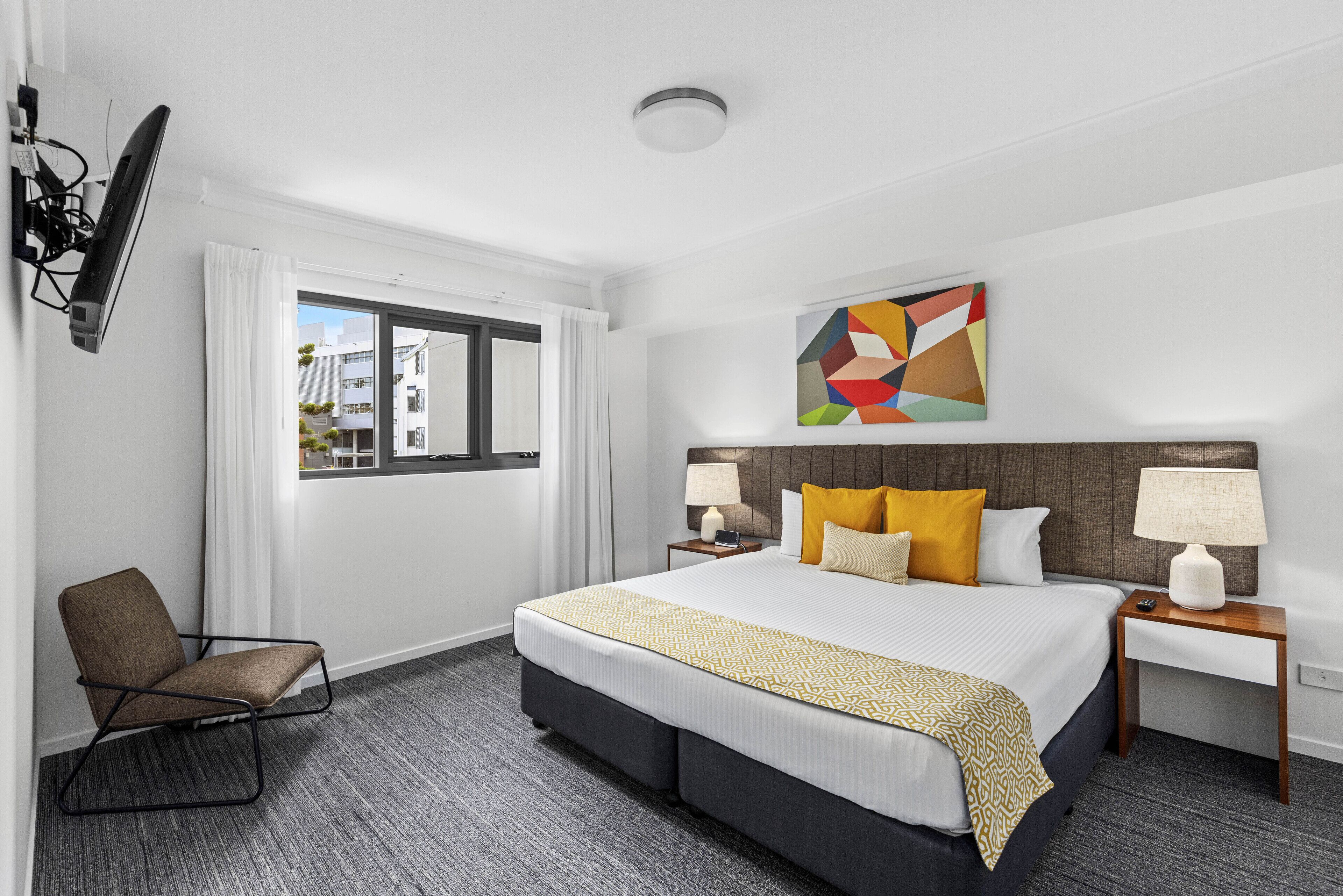 Una habitación de hotel con una cama grande, una silla, un televisor montado en la pared, una ventana con cortinas y un cuadro abstracto y colorido sobre la cama.