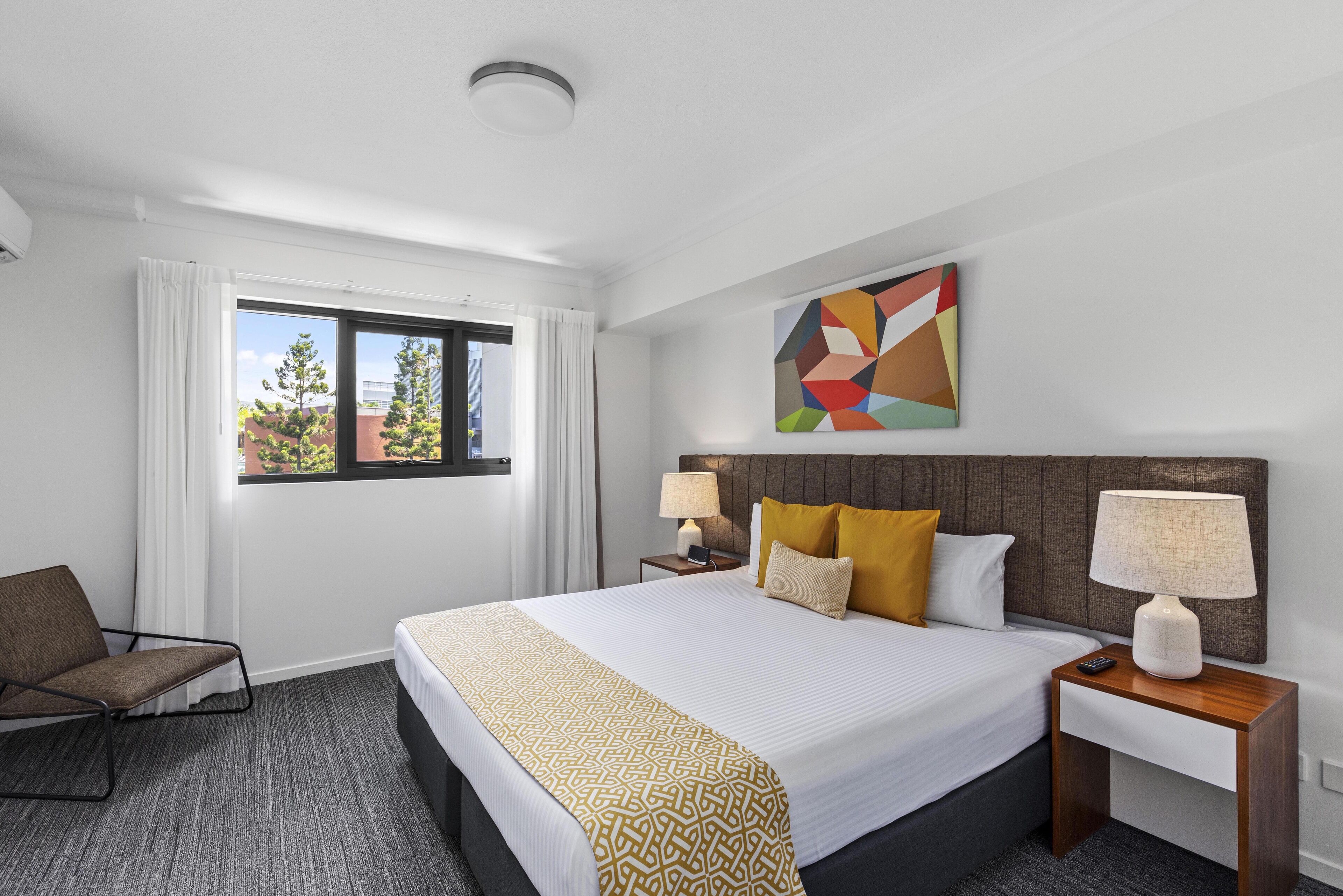 Een hotelkamer met een groot bed, twee nachtkastjes met lampen, een stoel, een raam met gordijnen en een kleurrijk abstract schilderij aan de muur.