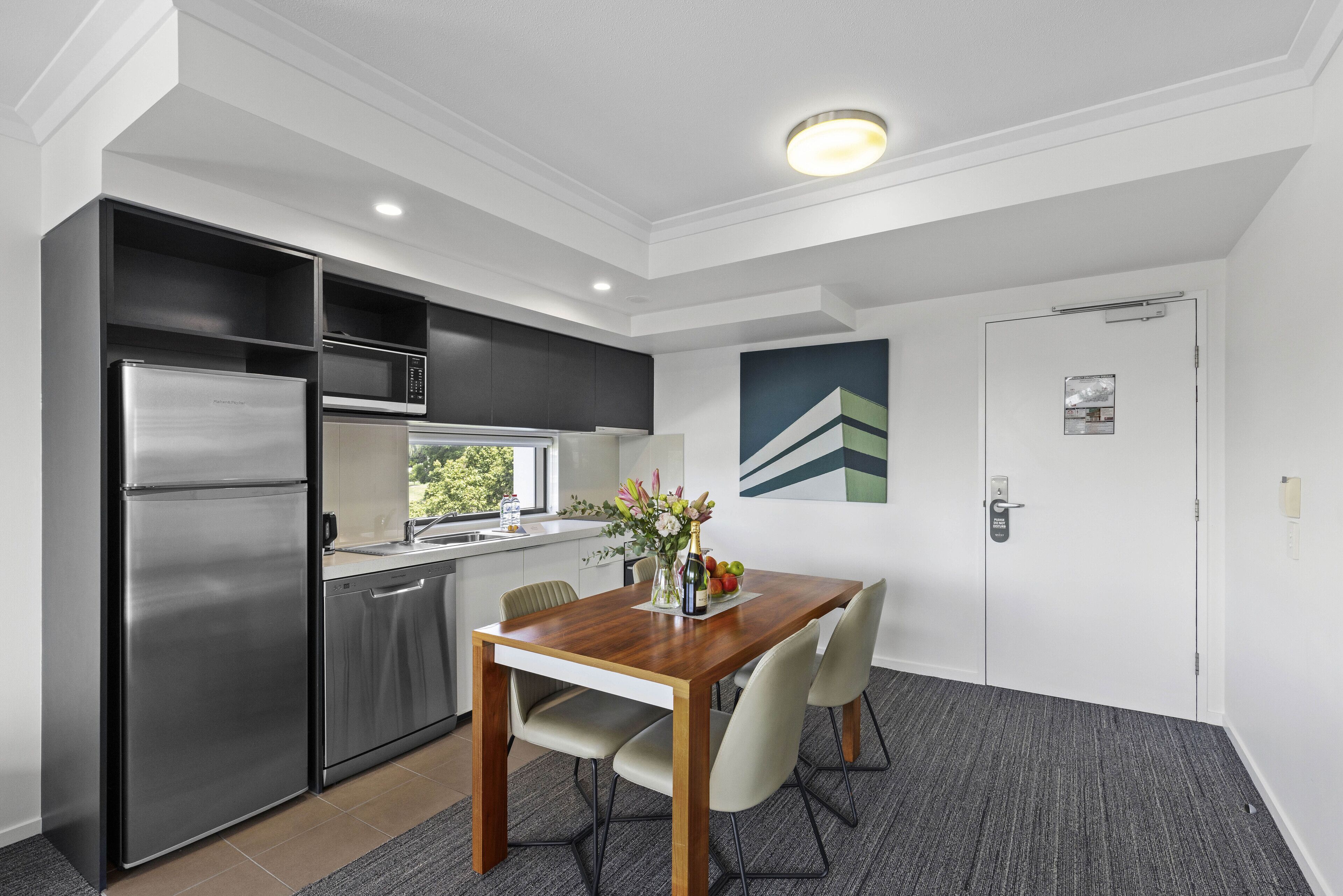 A modern kitchen with a stainless steel refrigerator, microwave, and dishwasher, a wooden dining table with chairs, and a door with a security lock.