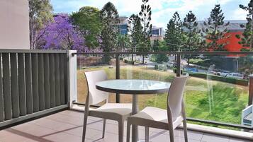 A balcony with a glass table and two chairs, overlooking a grassy area and buildings.