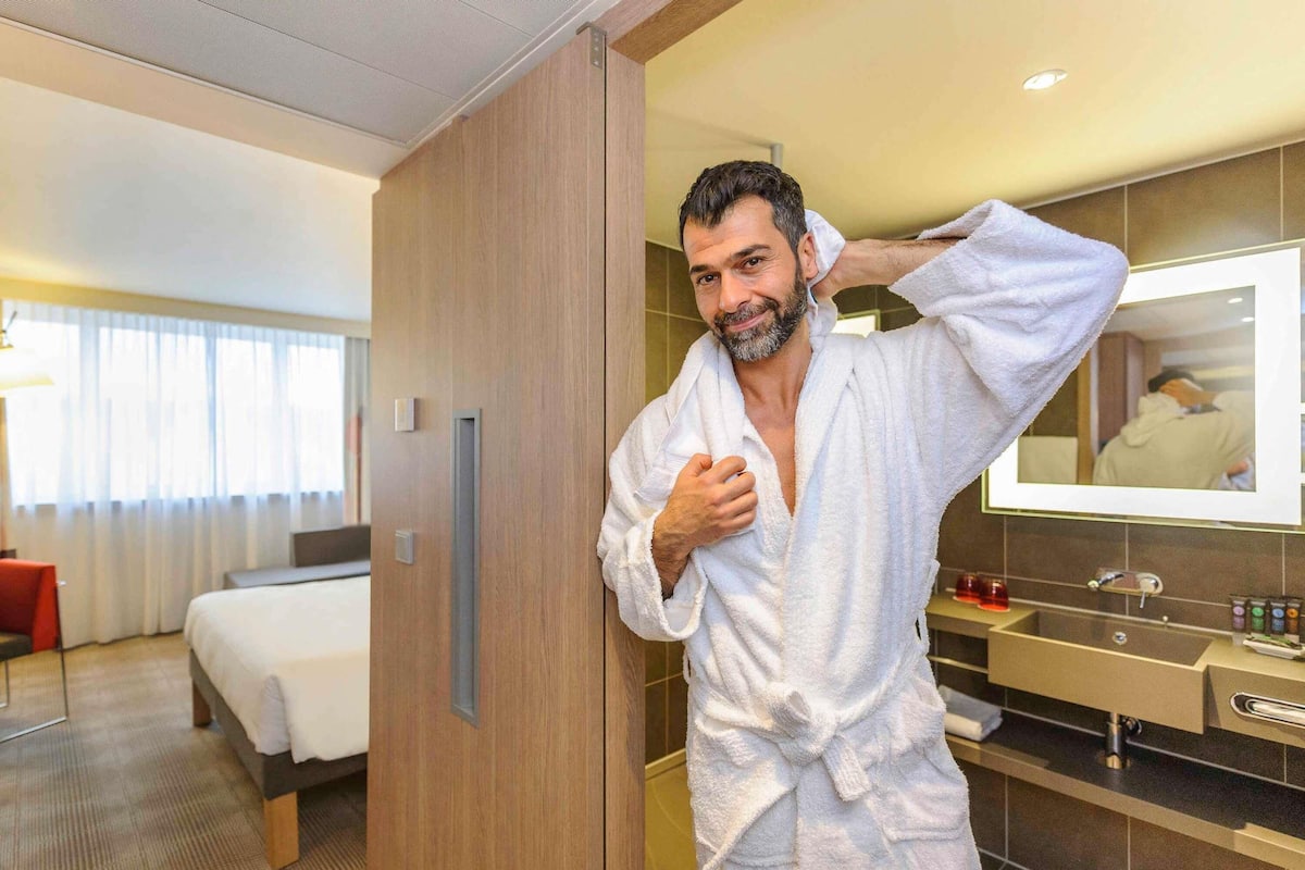 A hotel room with a bed, a wooden wardrobe, a bathroom with a mirror, and a red chair.