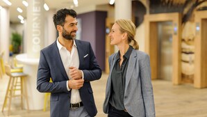 Un homme et une femme, vêtus de tenues professionnelles, sont en conversation dans un espace intérieur moderne, avec un bar en arrière-plan.