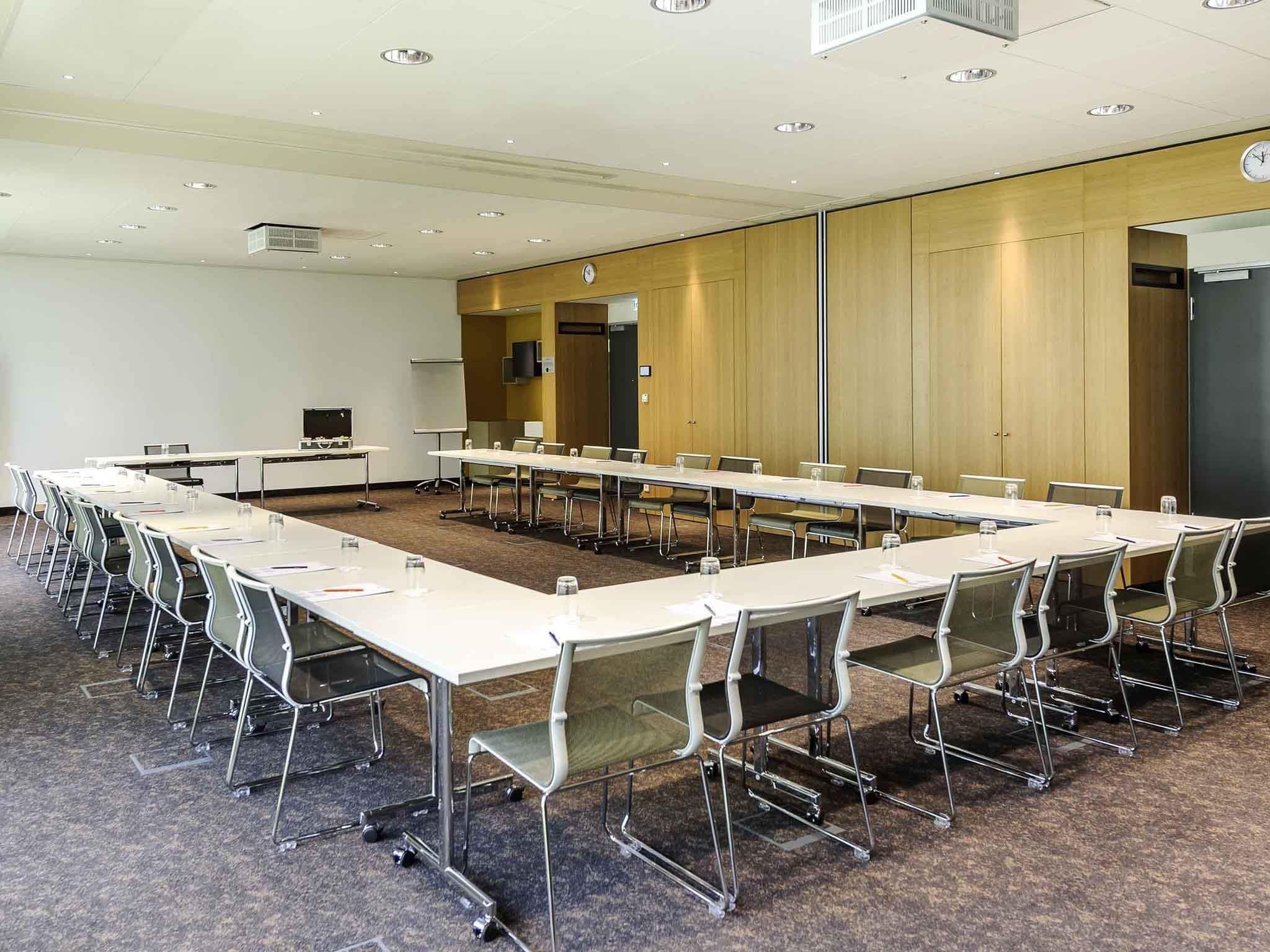 Uma sala de conferências com mesas e cadeiras compridas, um relógio na parede e um painel de parede de madeira.