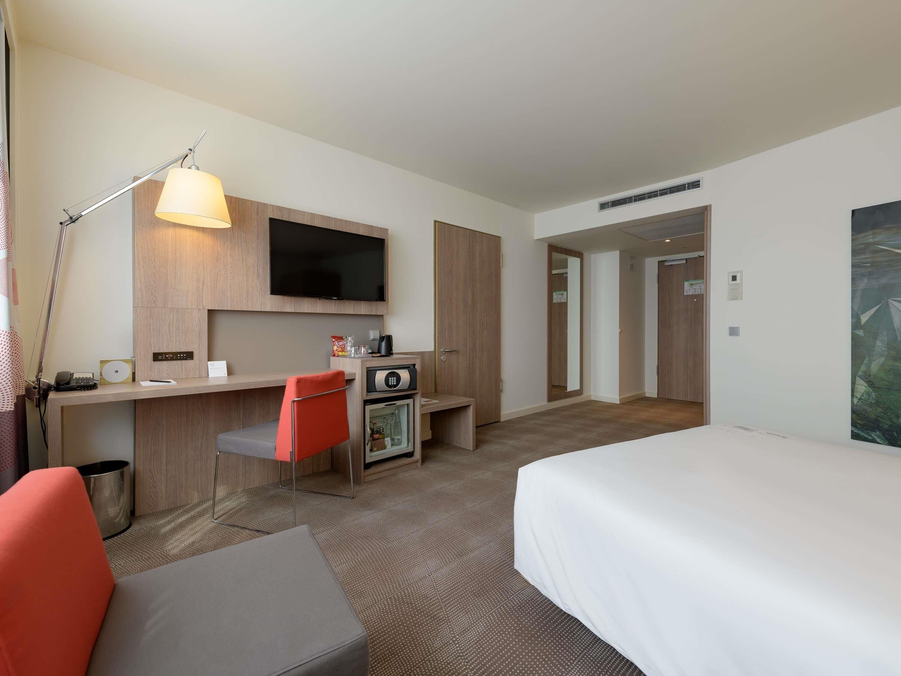 A hotel room with a bed, a desk with a flat-screen TV, a red chair, a small refrigerator, and a trash can.