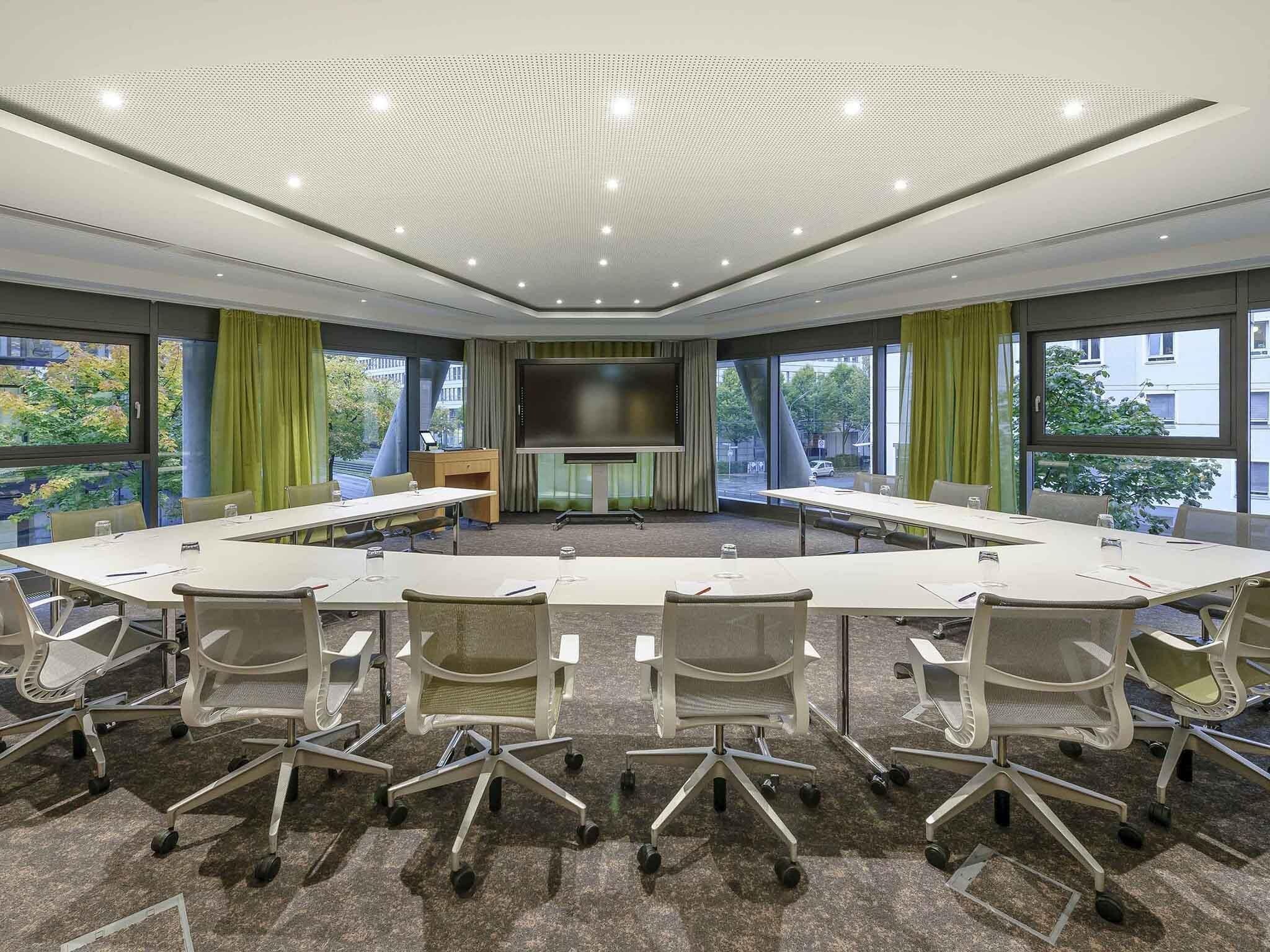Un salón de conferencias con una gran mesa, sillas, una pantalla y ventanas amplias.