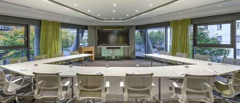 Un salón de conferencias con una gran mesa, sillas, una pantalla y ventanas amplias.