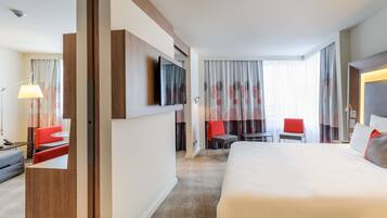 A hotel room with a bed, a desk, a chair, a TV mounted on the wall, and a window with curtains.